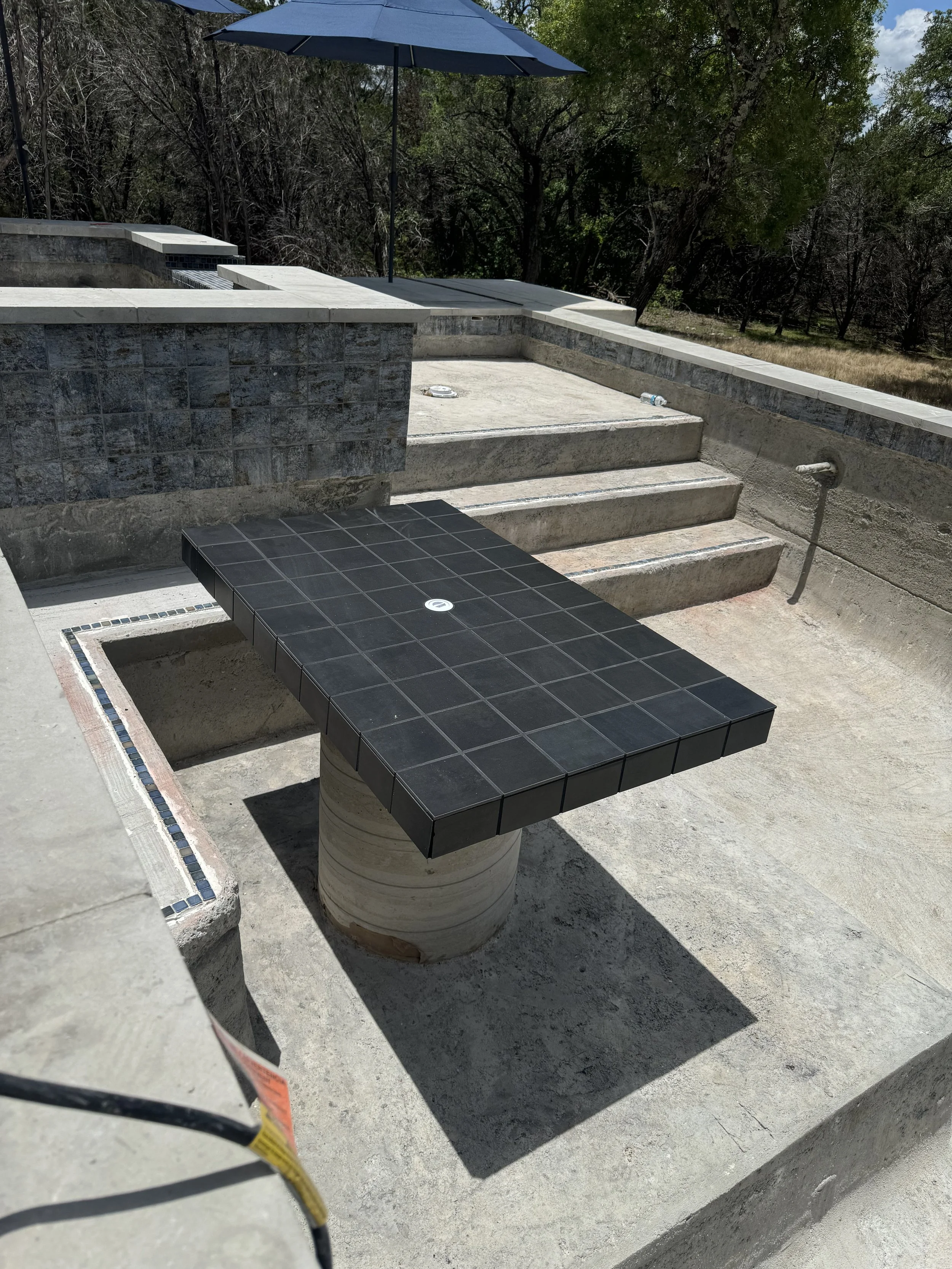 A black-tiled outdoor table with a circular concrete base, situated on a concrete surface near steps and a swimming pool, with a blue umbrella in the background.