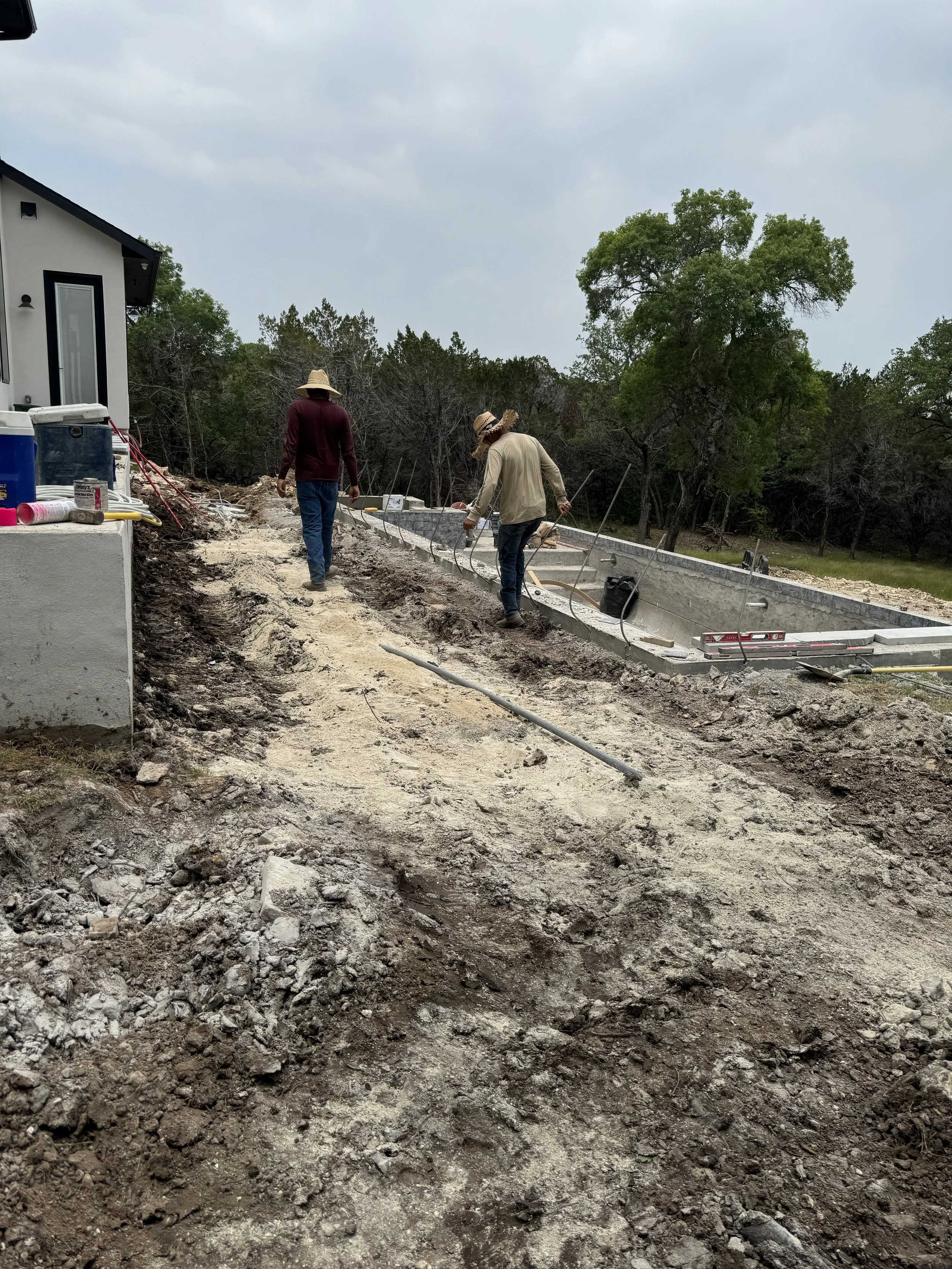 Two construction workers laying a concrete foundation for a building outdoors on a dirt site with trees in the background.