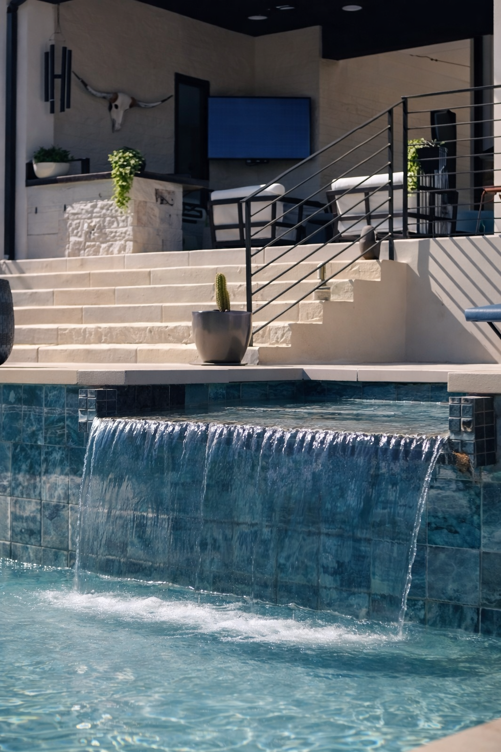 A swimming pool with a water feature spilling into the pool, surrounded by patio steps and outdoor furniture, with decorative plants and a mounted animal skull on the wall.