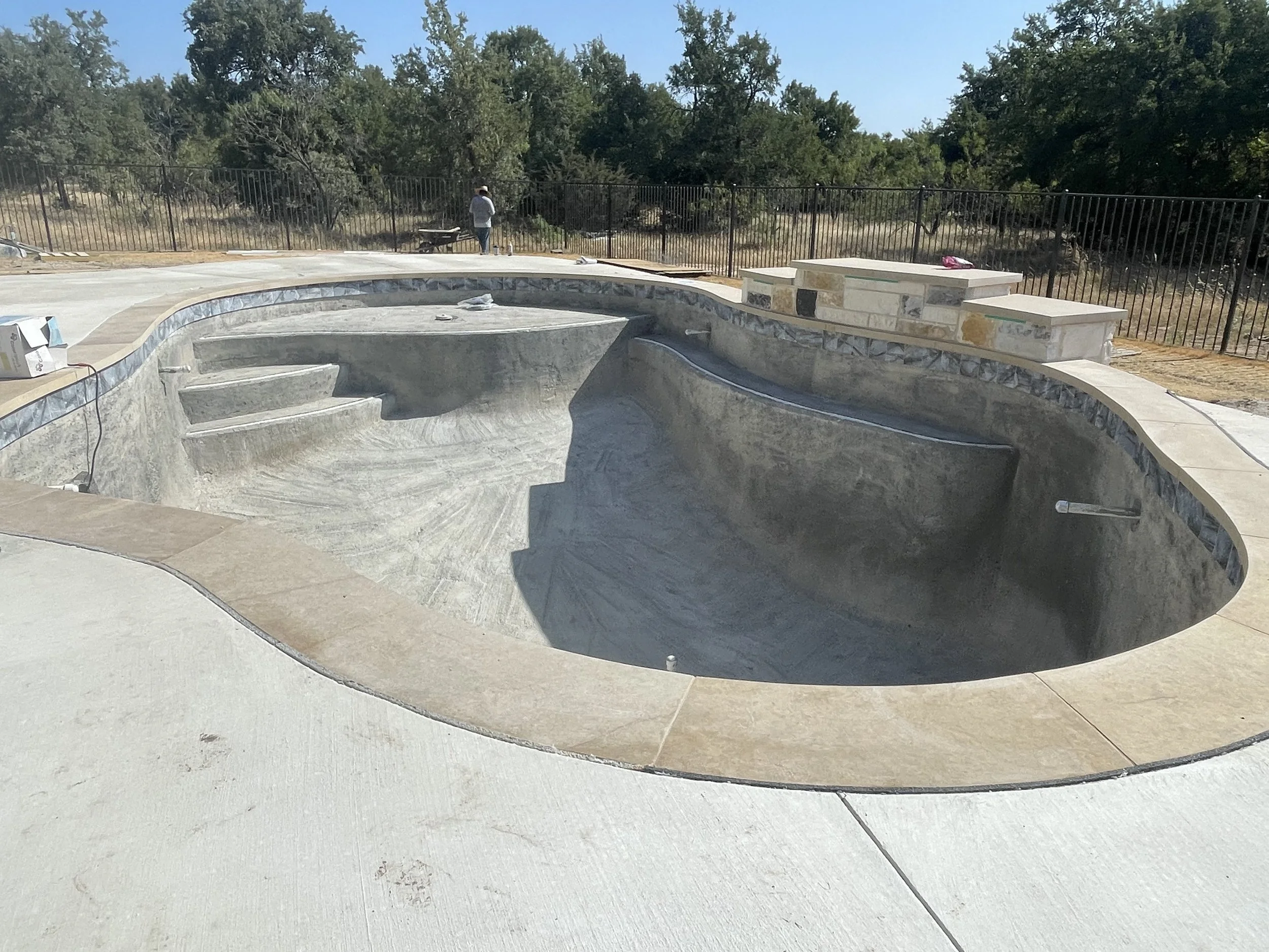 Empty swimming pool under construction surrounded by a beige stone patio, with a black metal fence and trees in the background.