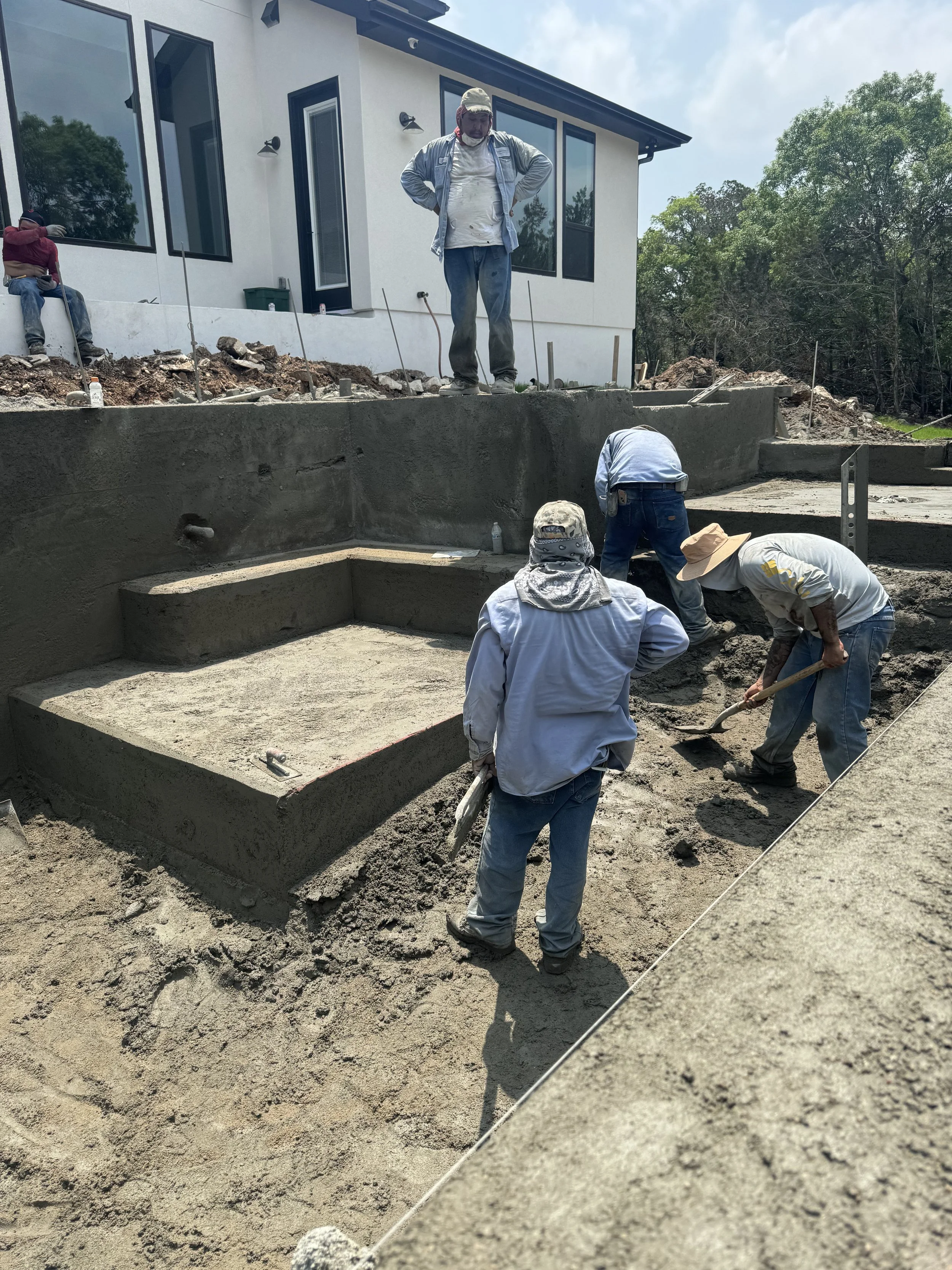 Construction workers are building a concrete foundation in a residential backyard, with a modern house in the background and trees on the side.