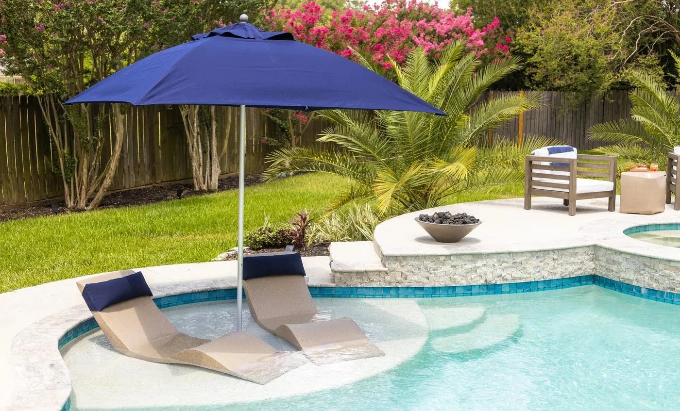 A backyard swimming pool area with two beige lounge chairs partially submerged in water, shaded by a blue umbrella, surrounded by green grass, trees, and pink flowering bushes, with outdoor seating in the background.