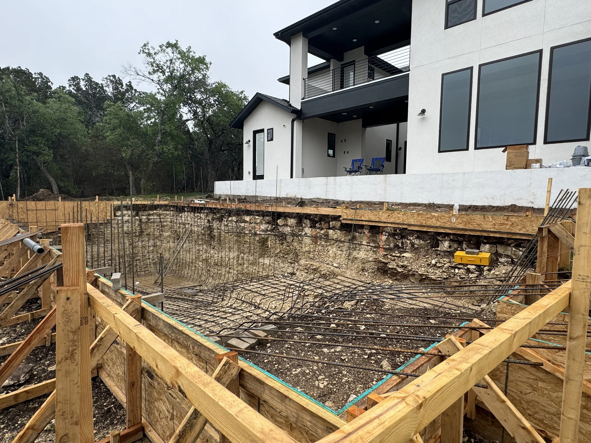 Construction site with a foundation trench and rebar for a new building, adjacent to a finished modern house with large windows and a balcony, in a wooded area.