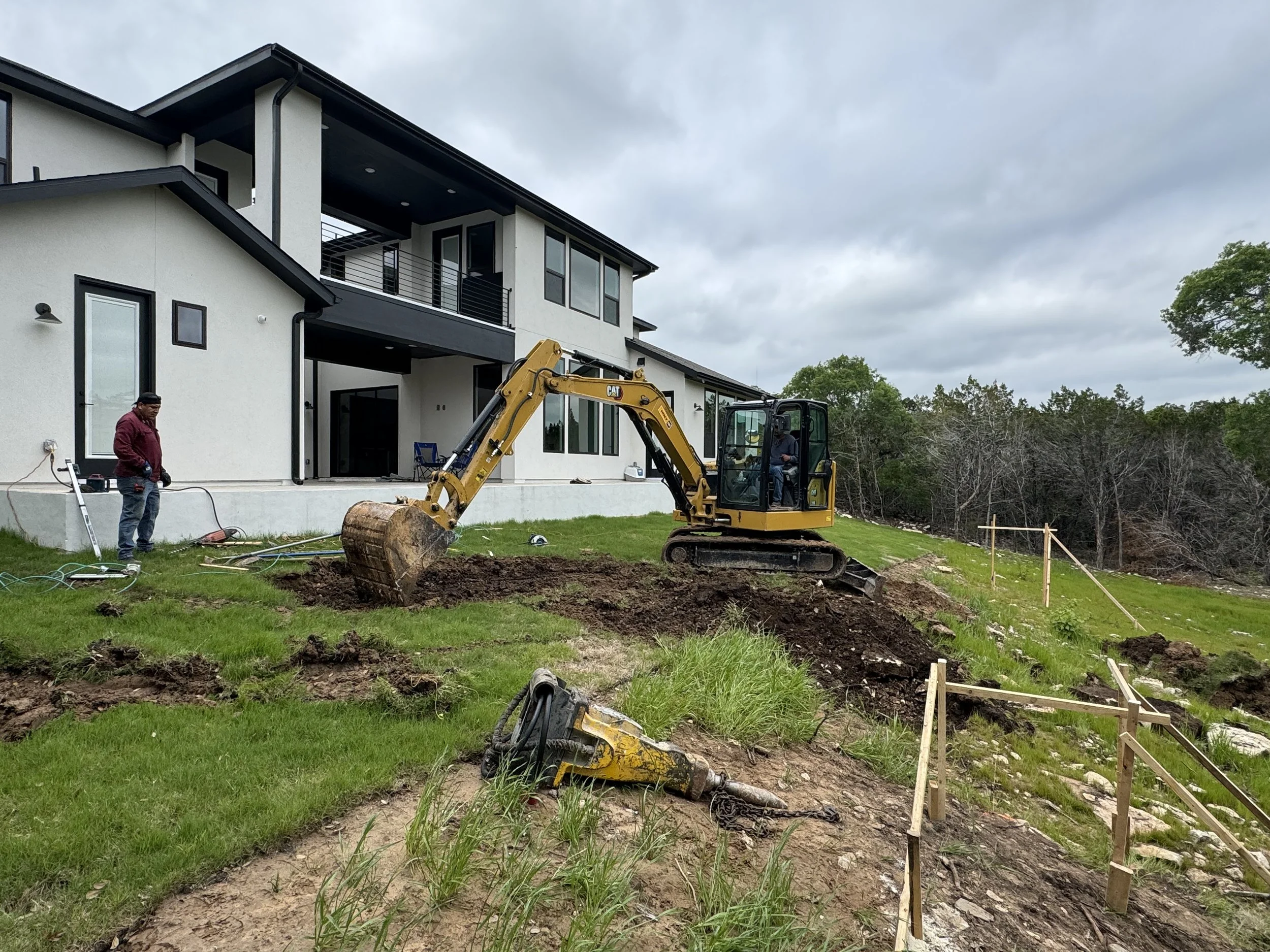 A construction site in a backyard with a small excavator digging into the ground near a modern two-story house. Two workers are present, one standing near the house and the other operating the excavator. Wooden stakes mark the area for excavation, wi