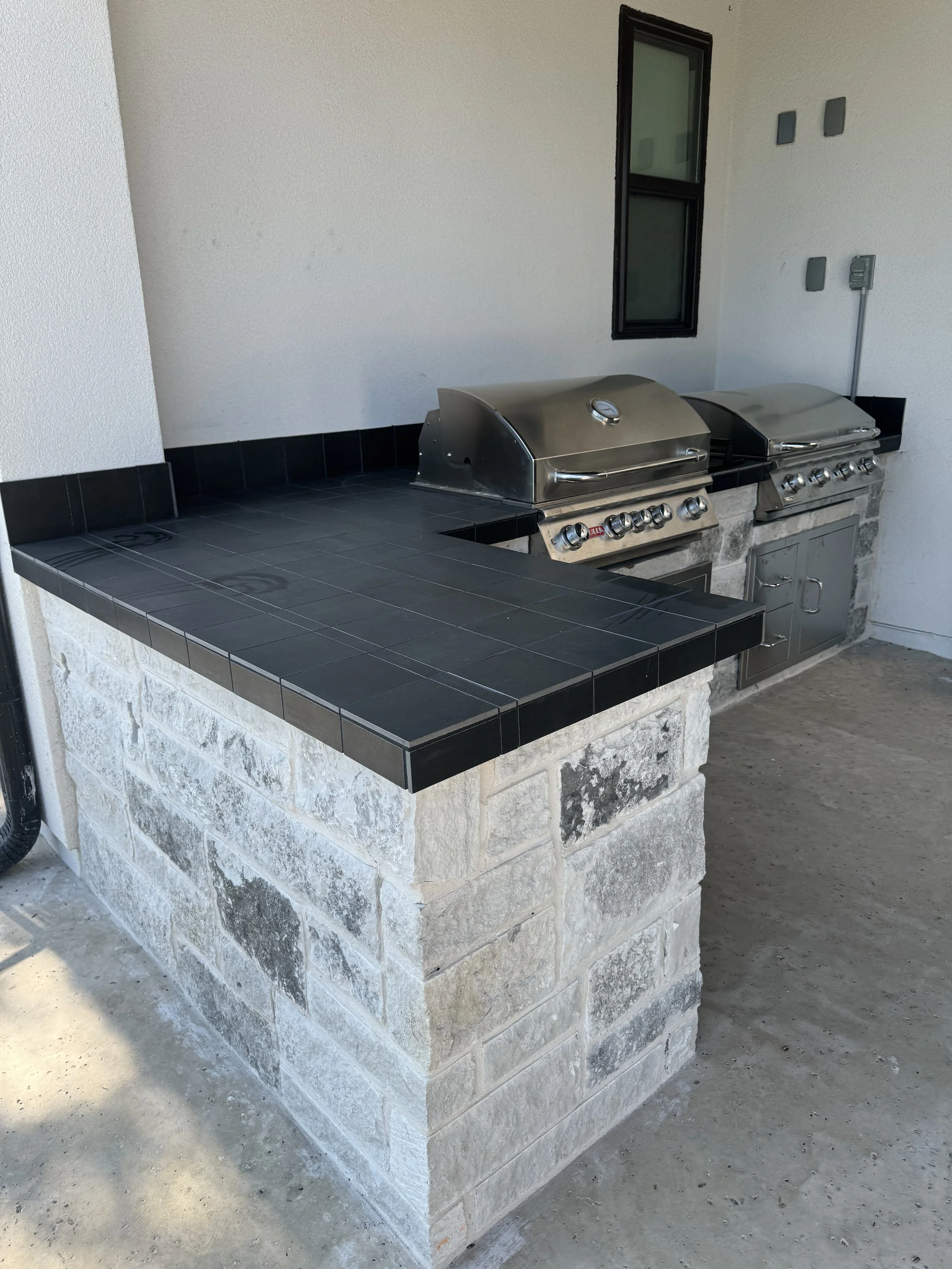 Outdoor barbecue area with two stainless steel grills, black tiled counter, and stone base, adjacent to a white wall with a small window.