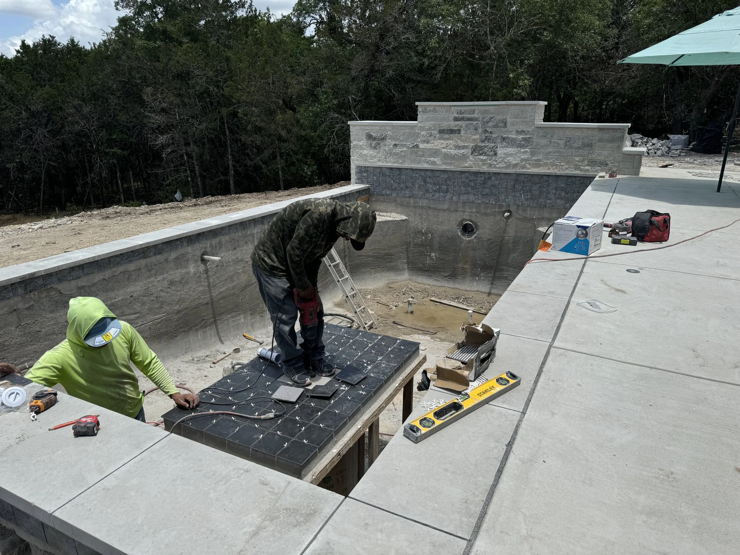 Two construction workers are working on the floor of a pool, installing tiles. One worker is wearing a bright green hoodie and the other is dressed in darker clothing with a camouflage hoodie. Construction tools and equipment are scattered around the