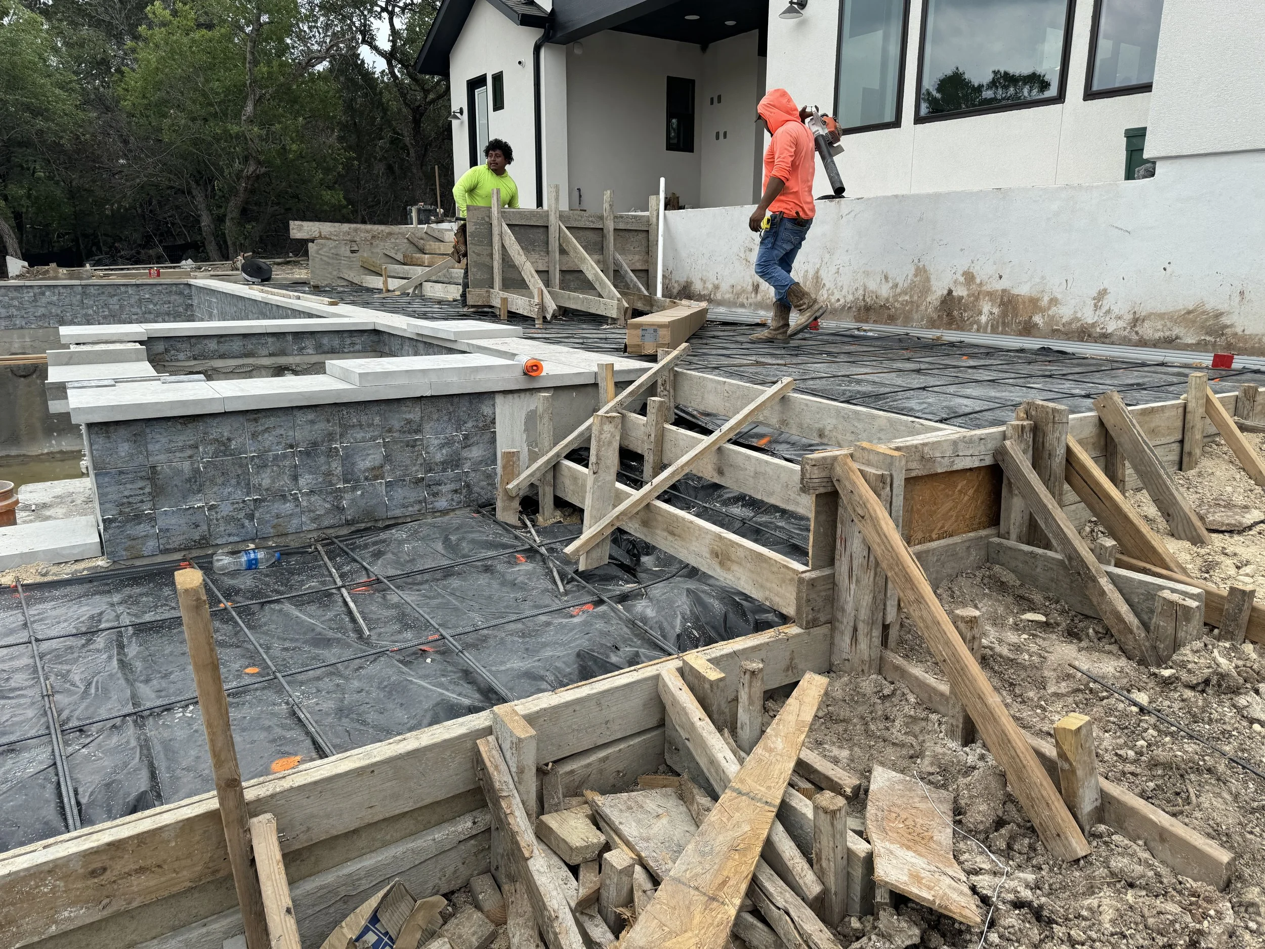 Construction workers pouring concrete on a building foundation at a house site, with plywood forms, rebar, and ongoing construction materials visible.
