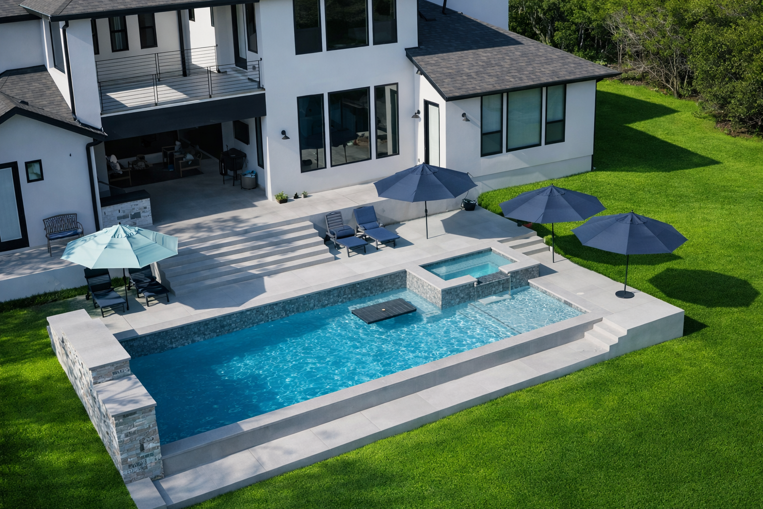 Aerial view of a modern backyard with a swimming pool, hot tub, and large patio with shaded umbrellas and lounge chairs adjacent to a white house surrounded by green grass and trees.
