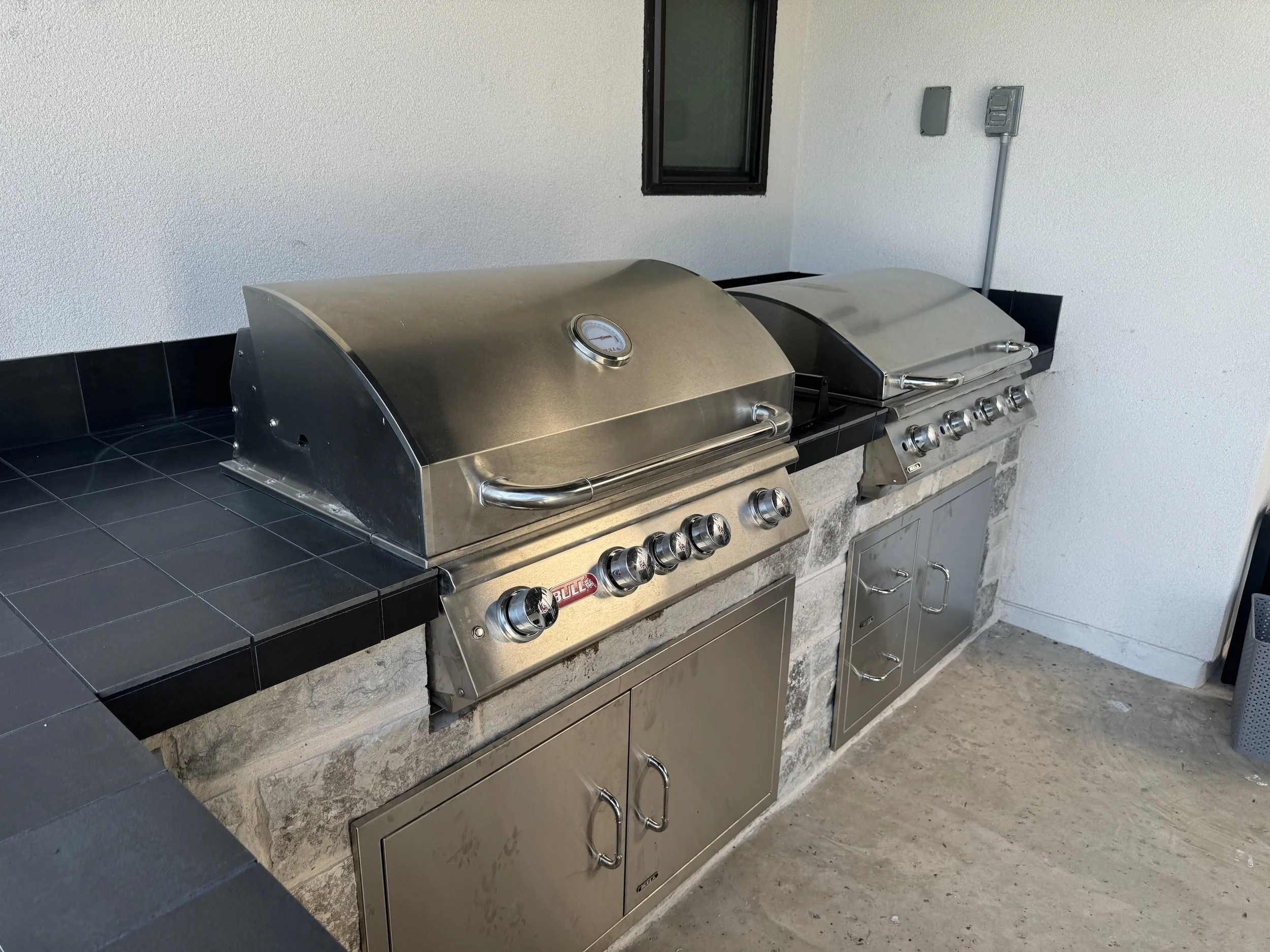 Two stainless steel grills on a black tiled countertop, with a white textured wall behind them, a small window, and utility switches on the wall.