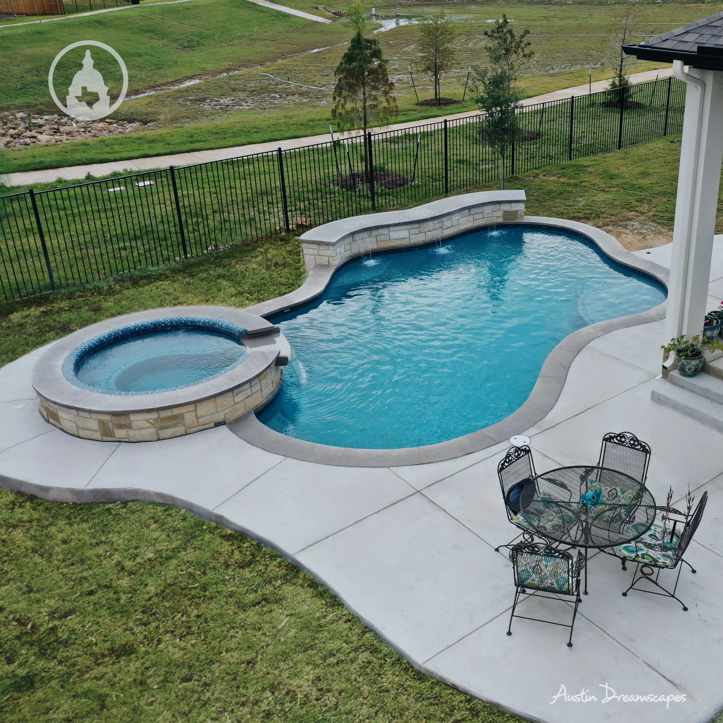 An outdoor swimming pool and hot tub on a concrete patio, with a grassy yard and a black metal fence in the background.