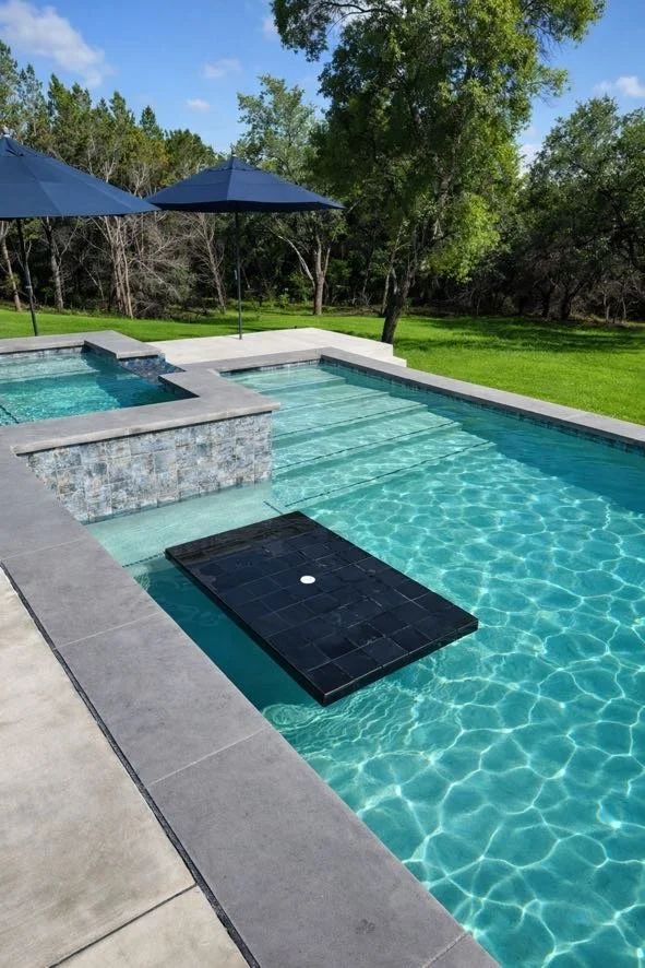 Outdoor swimming pool with a black floating platform, surrounded by a concrete deck and two blue umbrellas, with green grass and trees in the background on a sunny day.