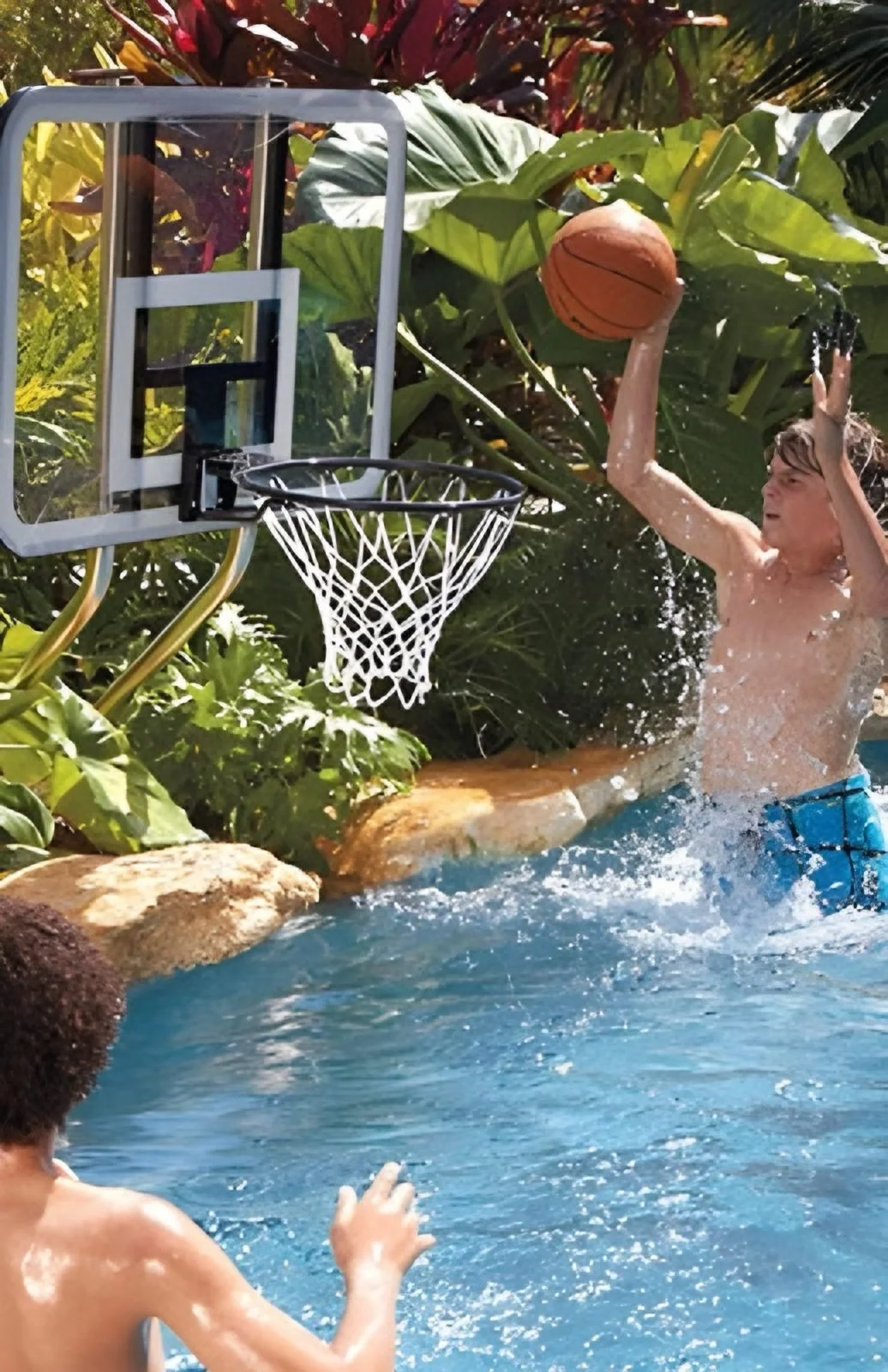 A boy in blue swim trunks playing water basketball in a pool, with another boy watching. The boy in the air is holding a basketball near a poolside basketball hoop surrounded by tropical plants.