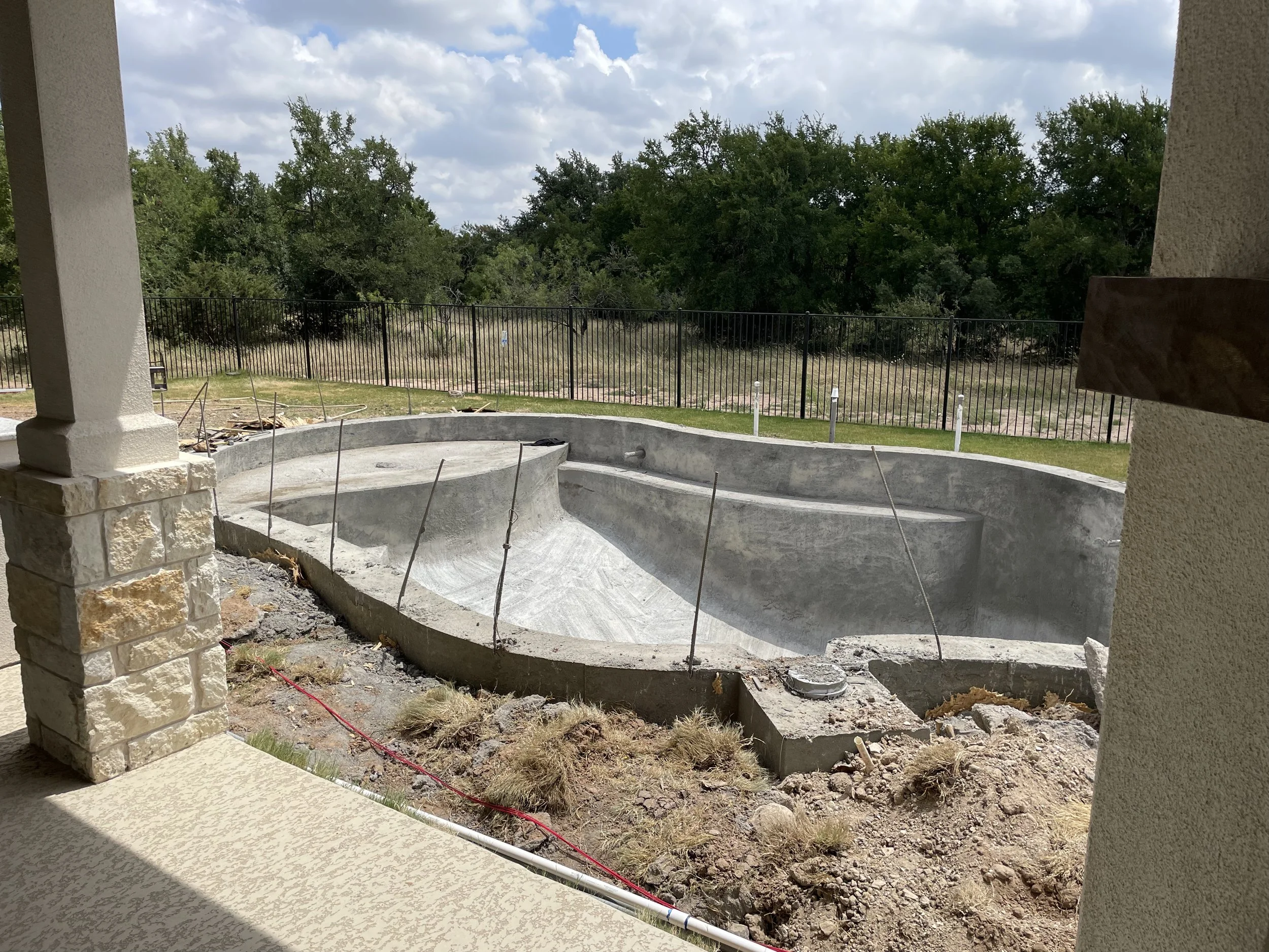 An empty residential swimming pool under construction, with concrete shell and exposed rebar, viewed from a covered patio, with a grassy yard and trees in the background.