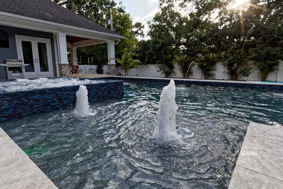 Modern backyard swimming pool with water jets and a hot tub, surrounded by trees and a white fence, in front of a house with a porch.