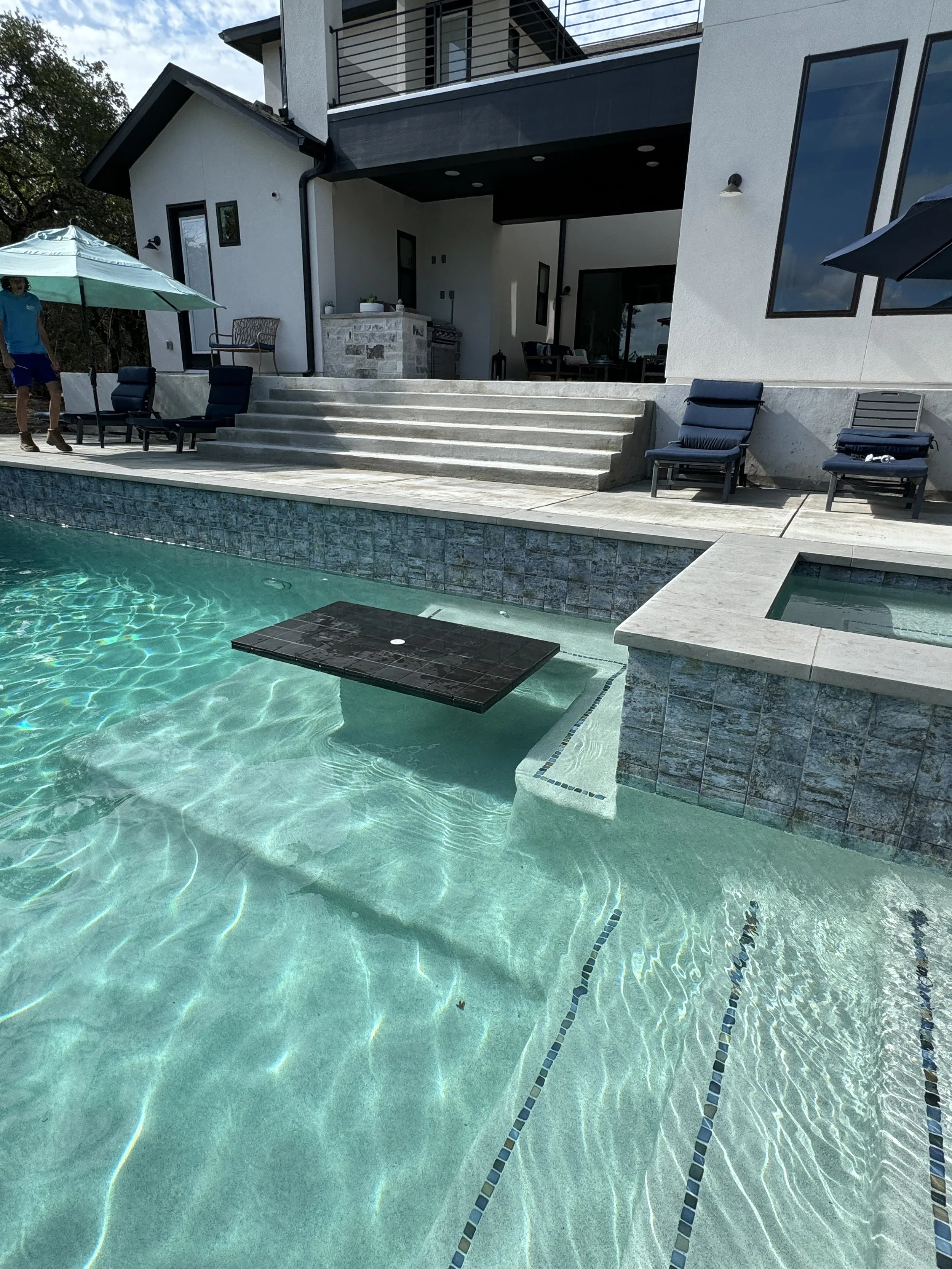 A modern backyard with a swimming pool in front of a white house with large windows and a patio area, featuring lounge chairs, an umbrella, and a person standing near the umbrellas.
