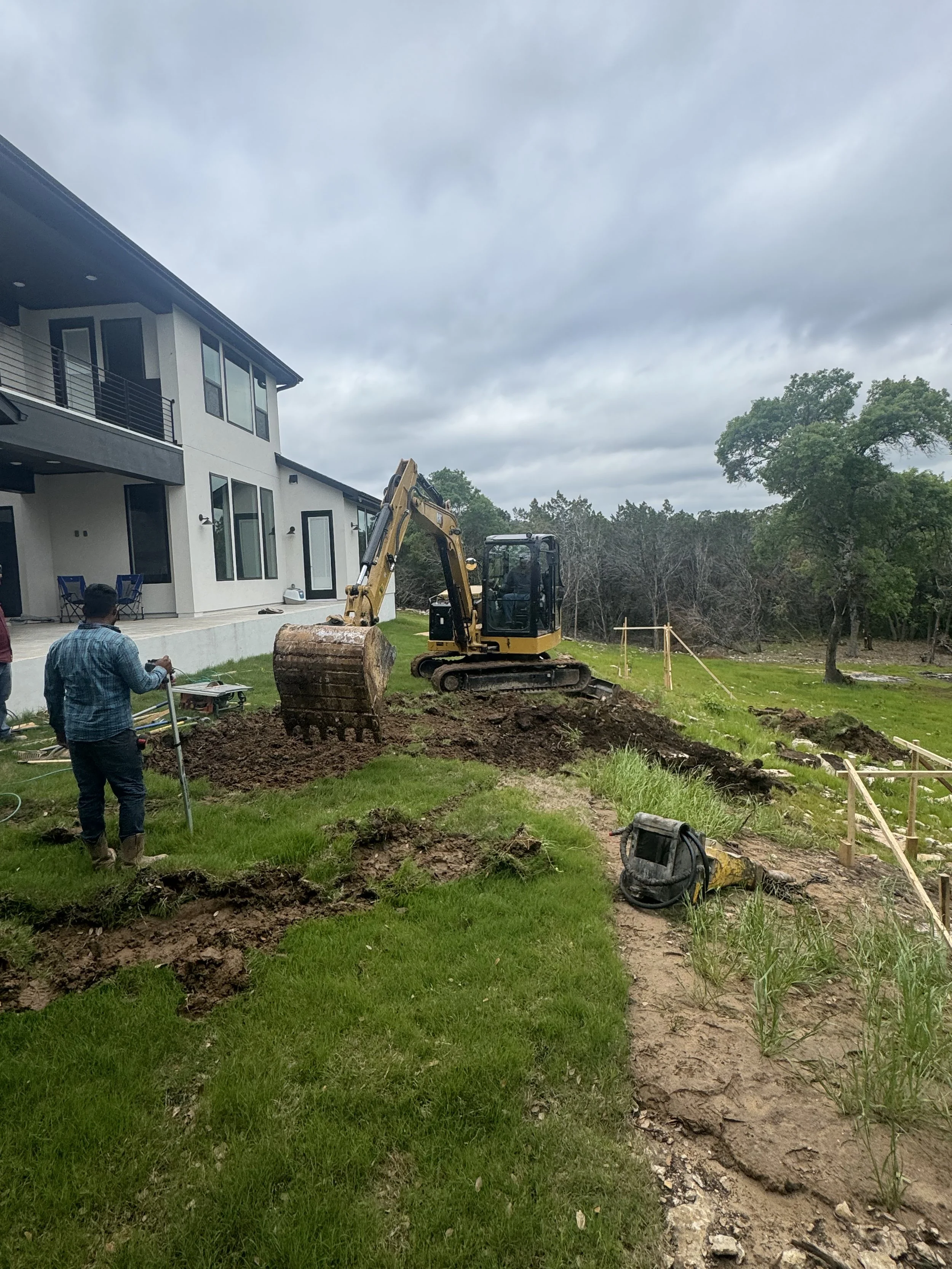 Construction site with a small excavator digging on a grassy area near a modern house, with workers and construction equipment present, cloudy sky overhead.