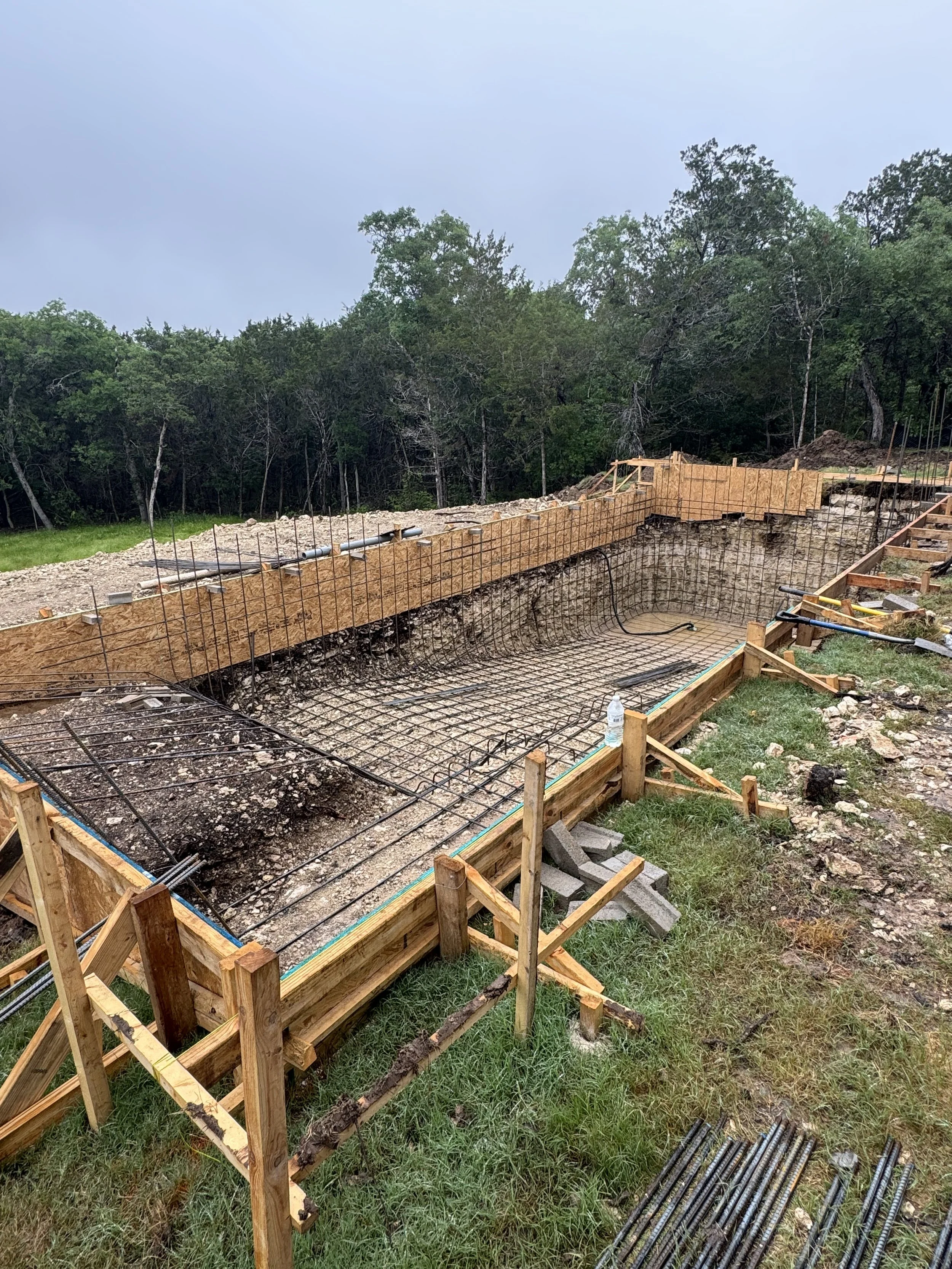 Construction site for a swimming pool with wooden forms and rebar framework in a backyard, surrounded by grass and trees in the background.