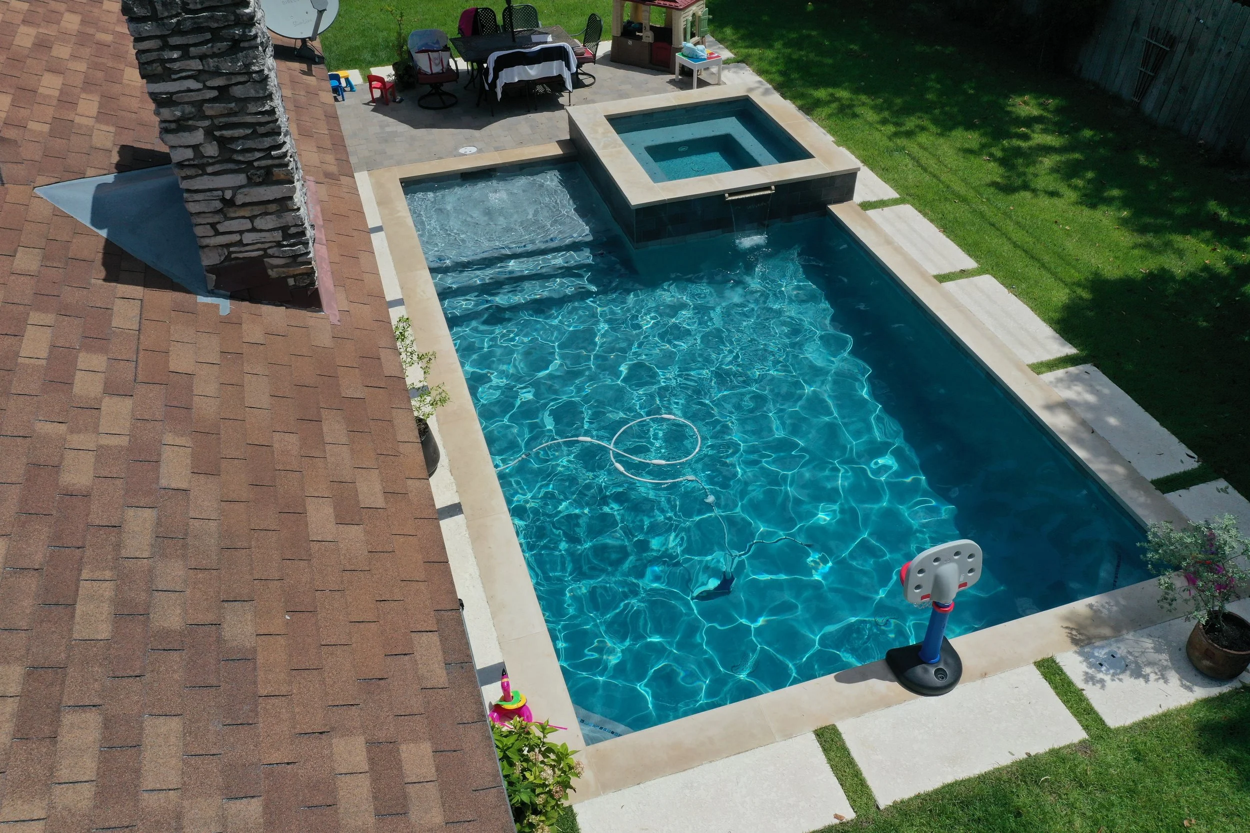 A backyard swimming pool with a small attached spa, a basketball hoop, a patio with outdoor furniture, and a grassy area with shade trees.