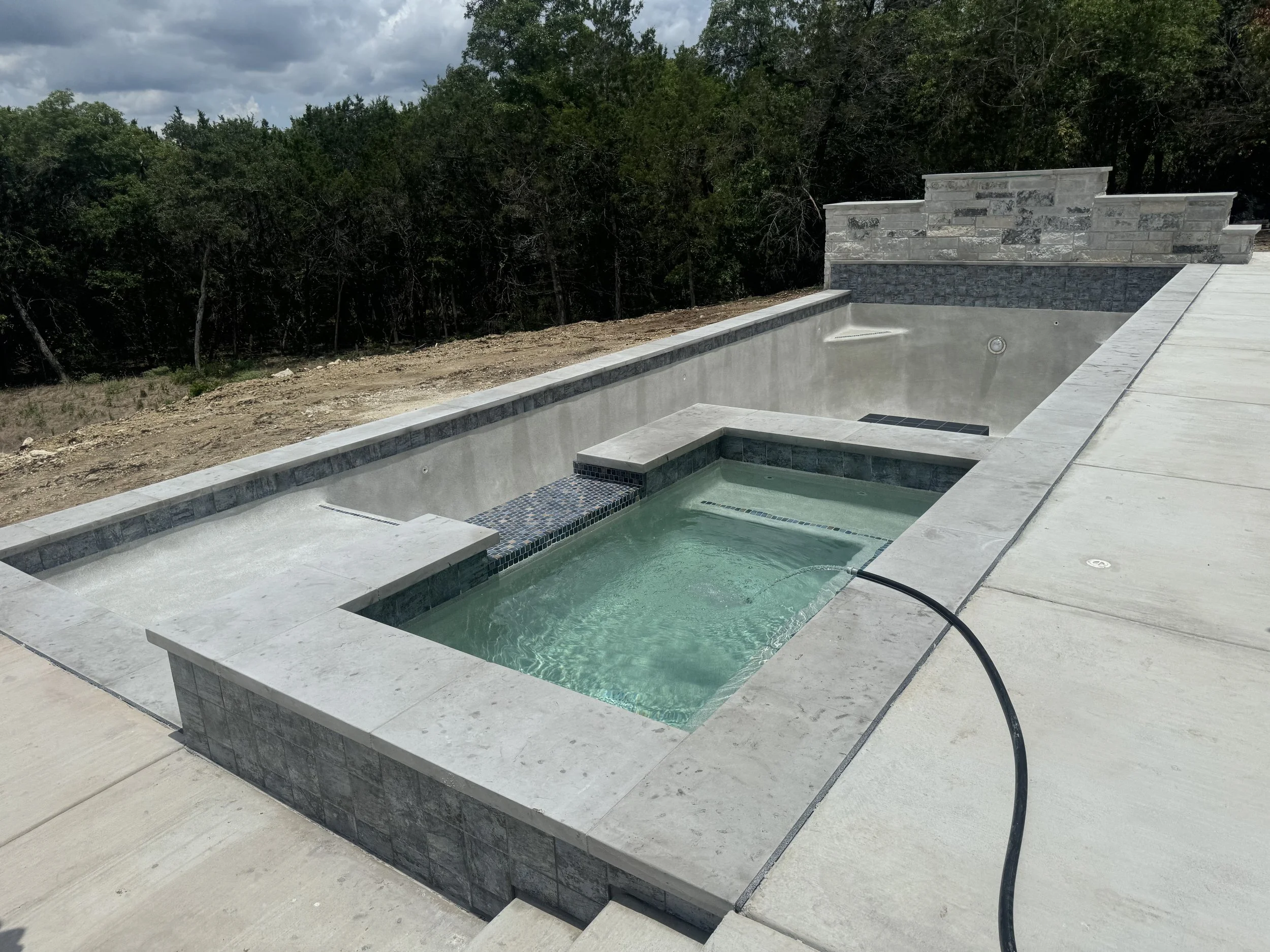 Empty in-ground swimming pool with hot tub, under construction, with a hose filling the pool, outdoors with trees in the background.