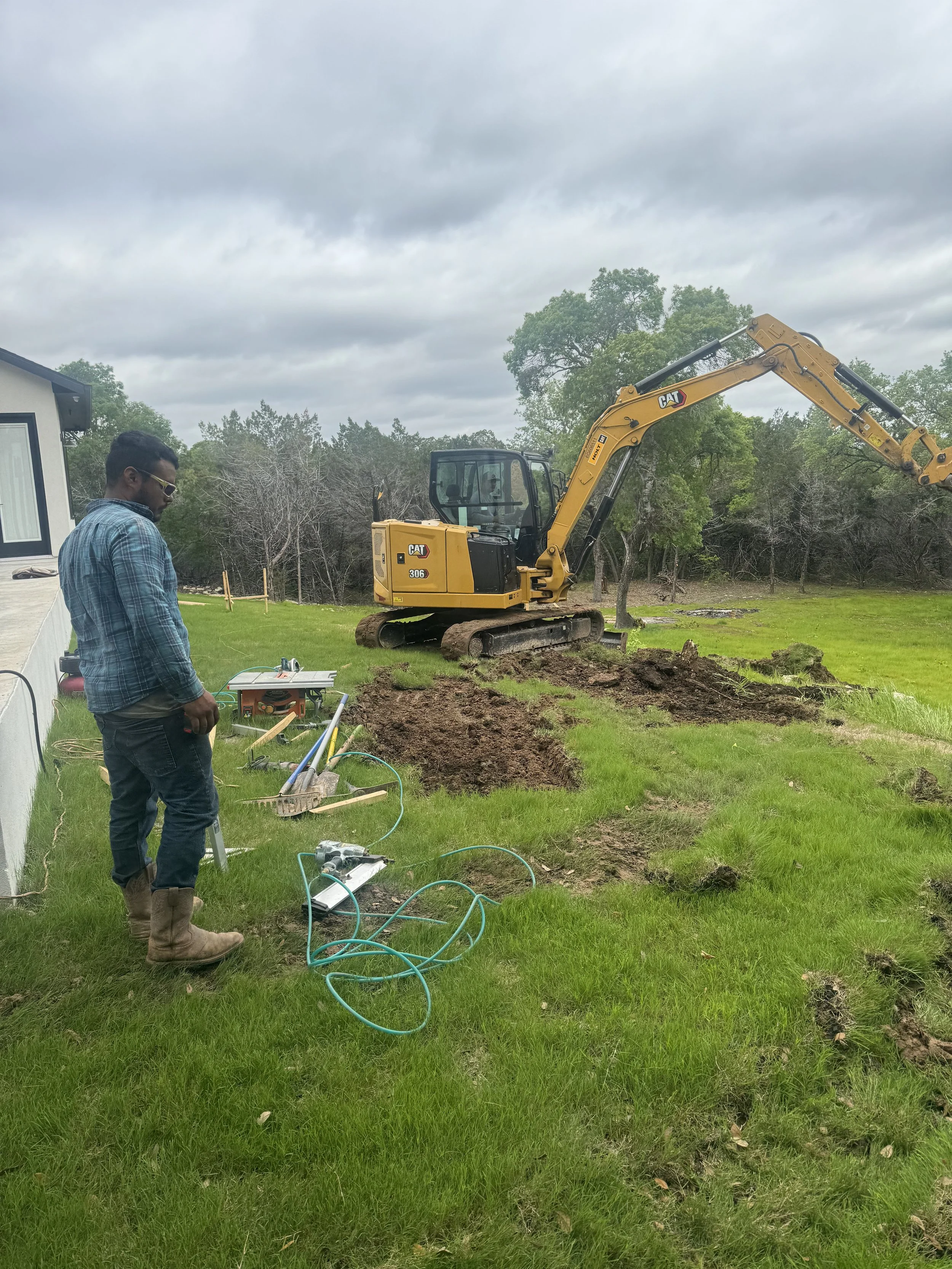A construction worker and a mini excavator working on a backyard landscape project, with tools and cables on the grass under an overcast sky.
