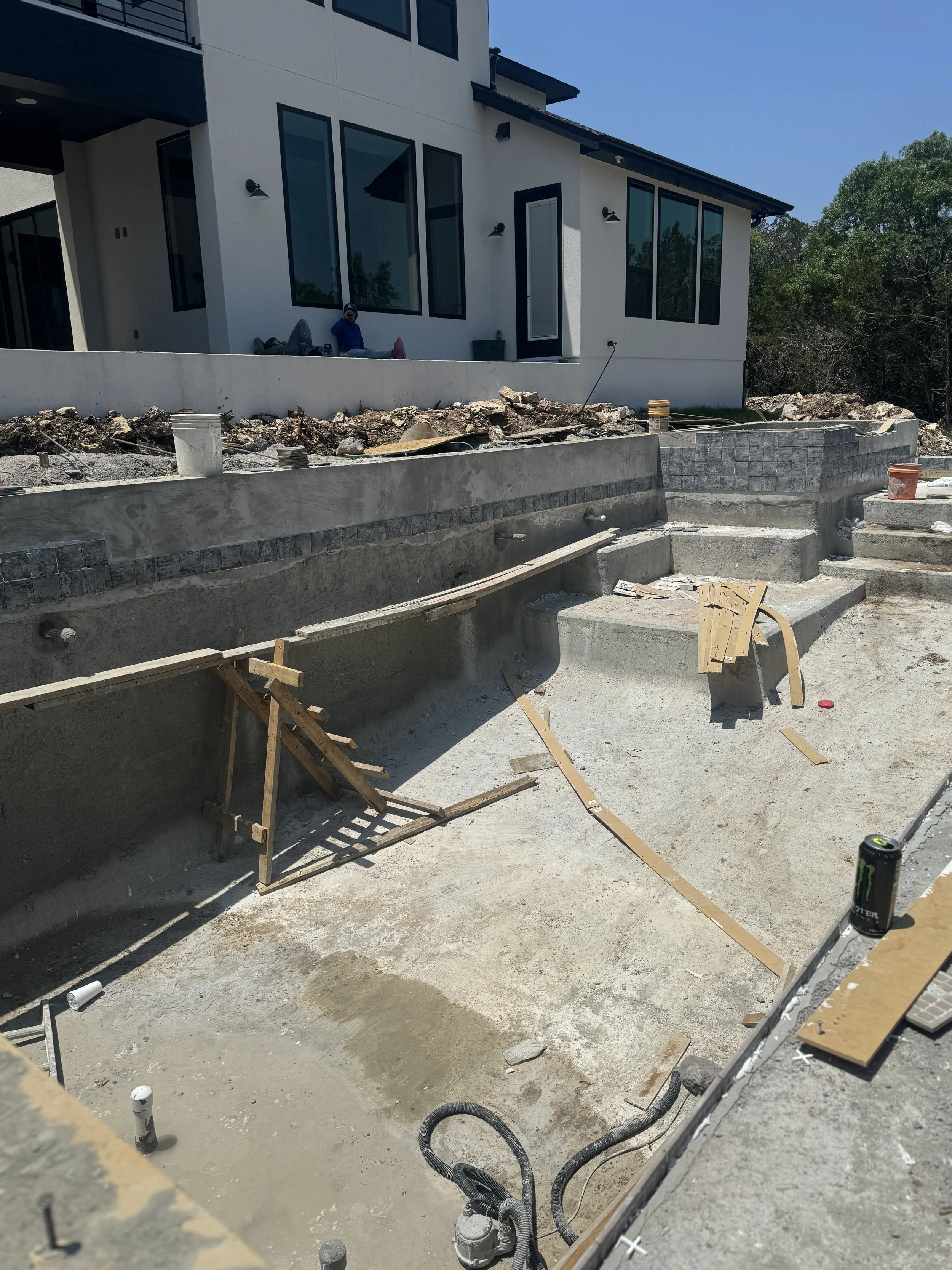 Construction site in front of a modern white house with large windows, showing ongoing building work for a pool or outdoor area.