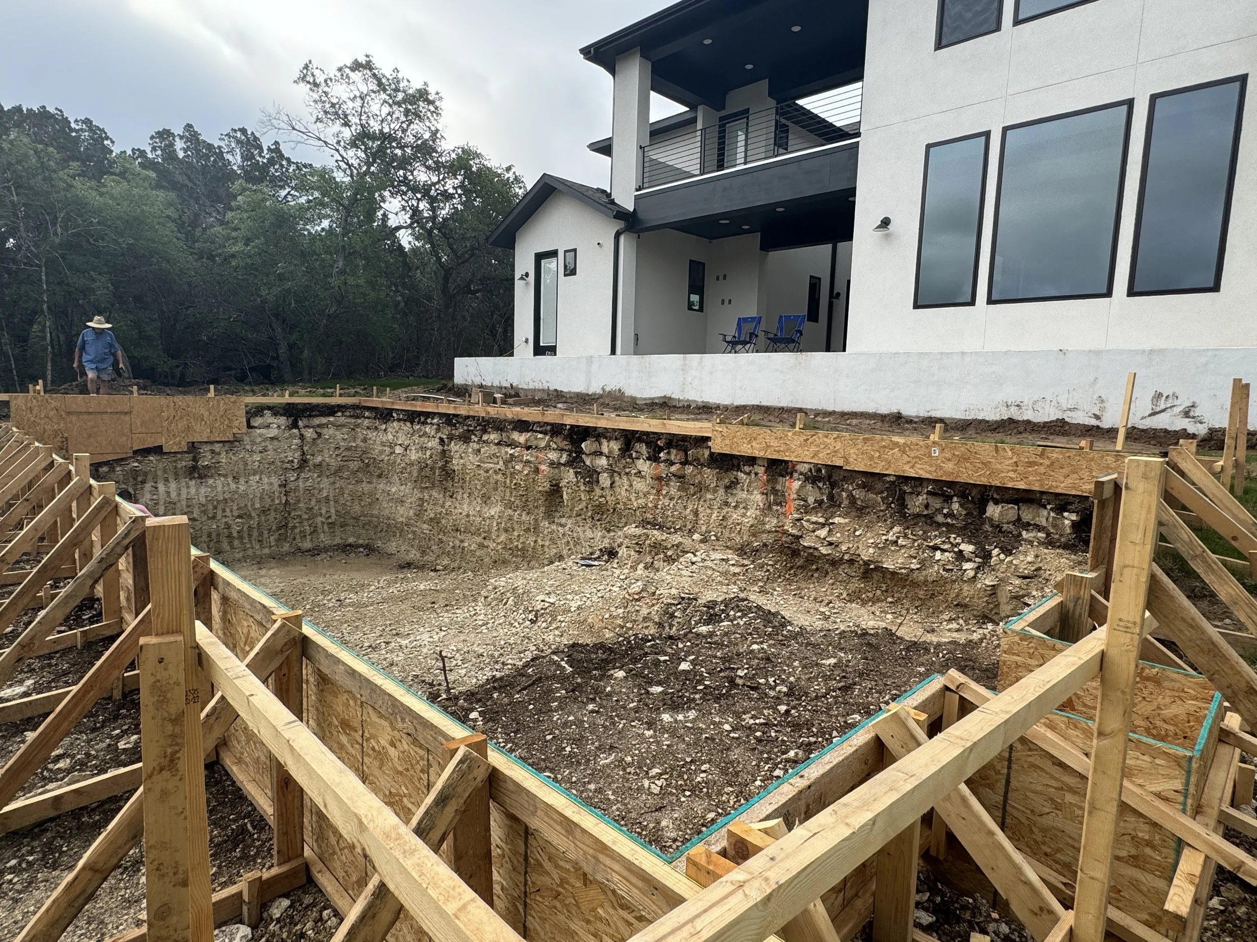 Construction site with a house in the background and an excavated area in the foreground framed with wooden formwork for a foundation or pool.