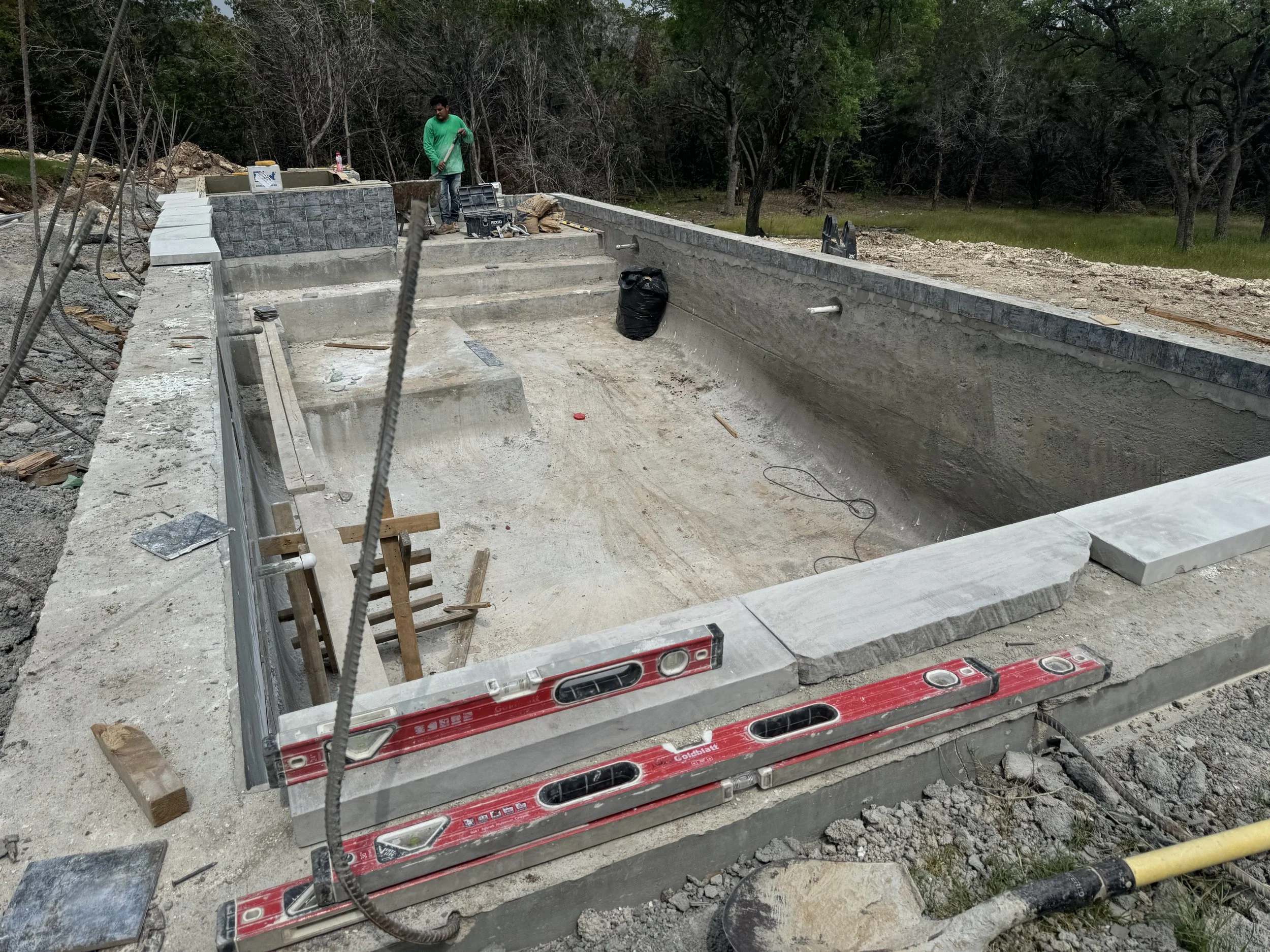 Under construction swimming pool with workers, concrete work in progress, surrounded by trees.