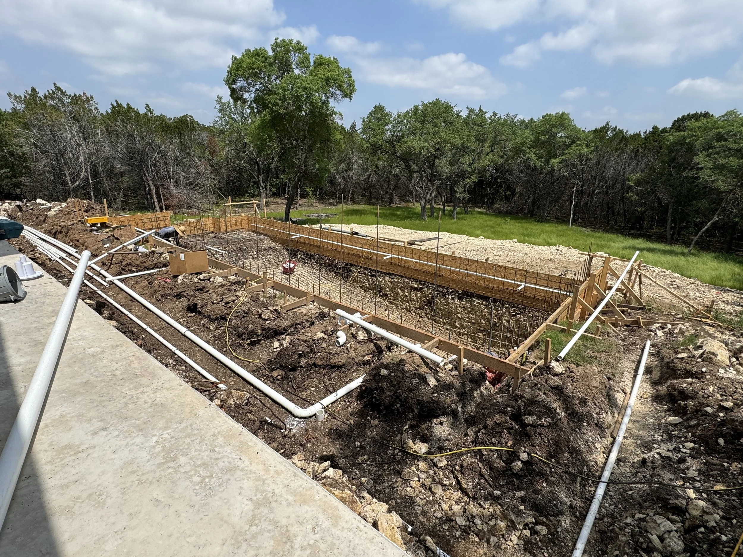 Construction site for a building foundation with wooden forms and rebar, surrounded by dirt, pipes, and tools, with trees and blue sky in the background.