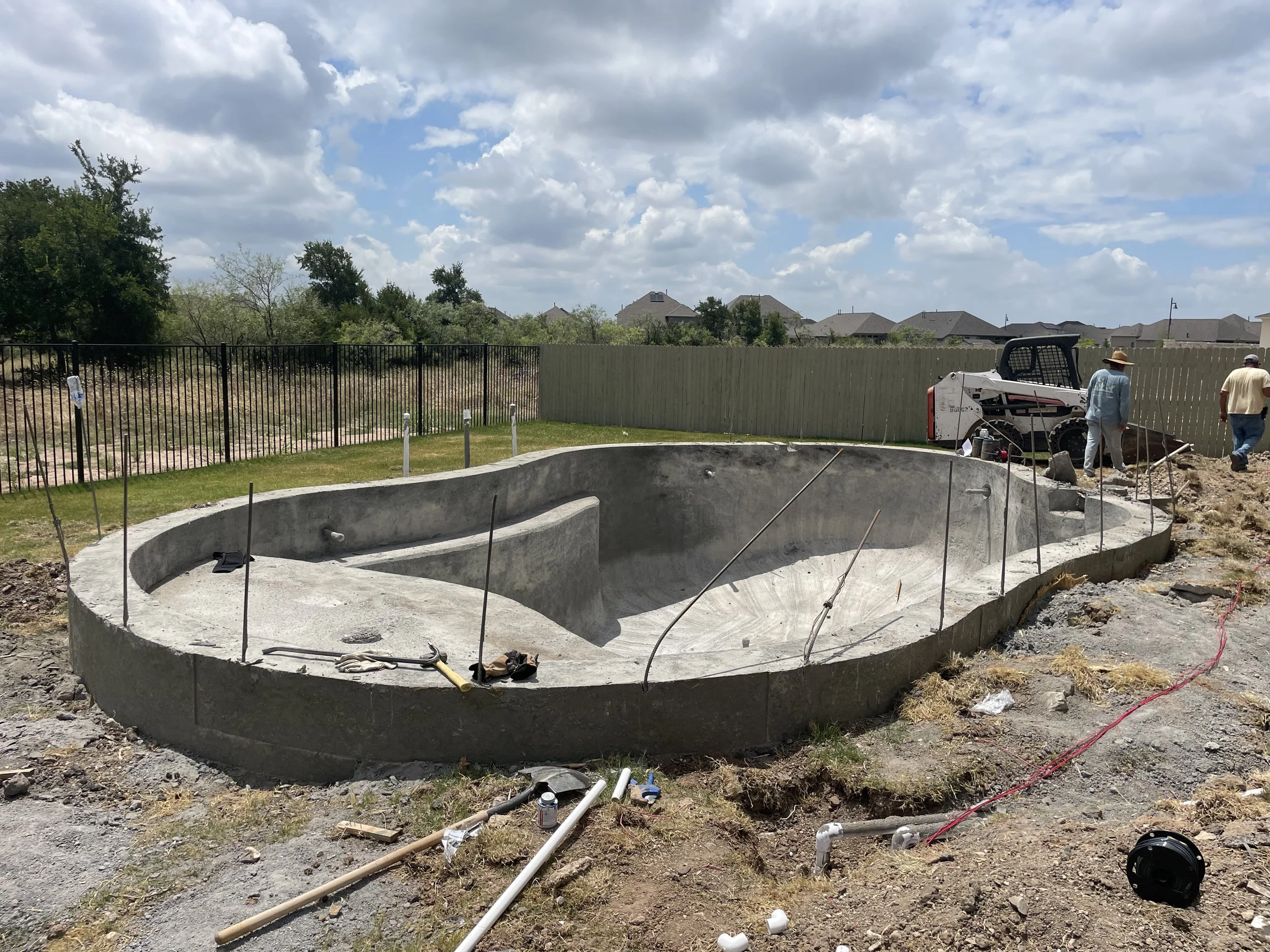 Incomplete in-ground swimming pool under construction with workers and construction equipment nearby, surrounded by a fenced yard.