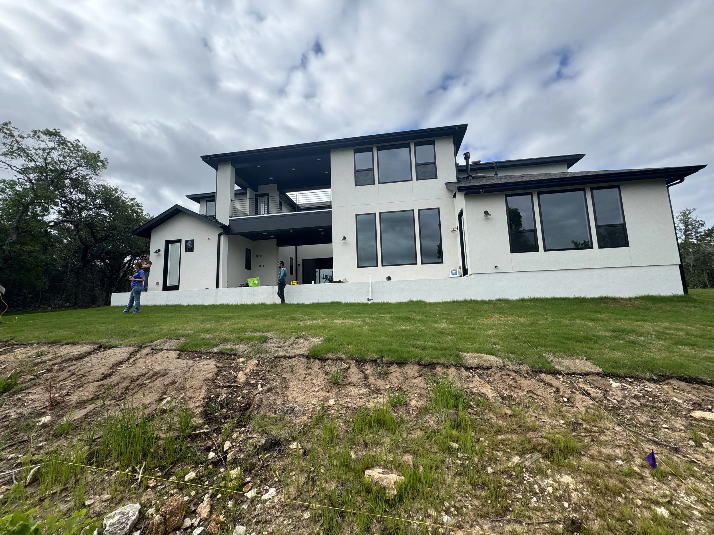 Modern two-story house with large windows and a white exterior, situated on a grassy hill with some trees in the background, under a cloudy sky, with three people standing outside.