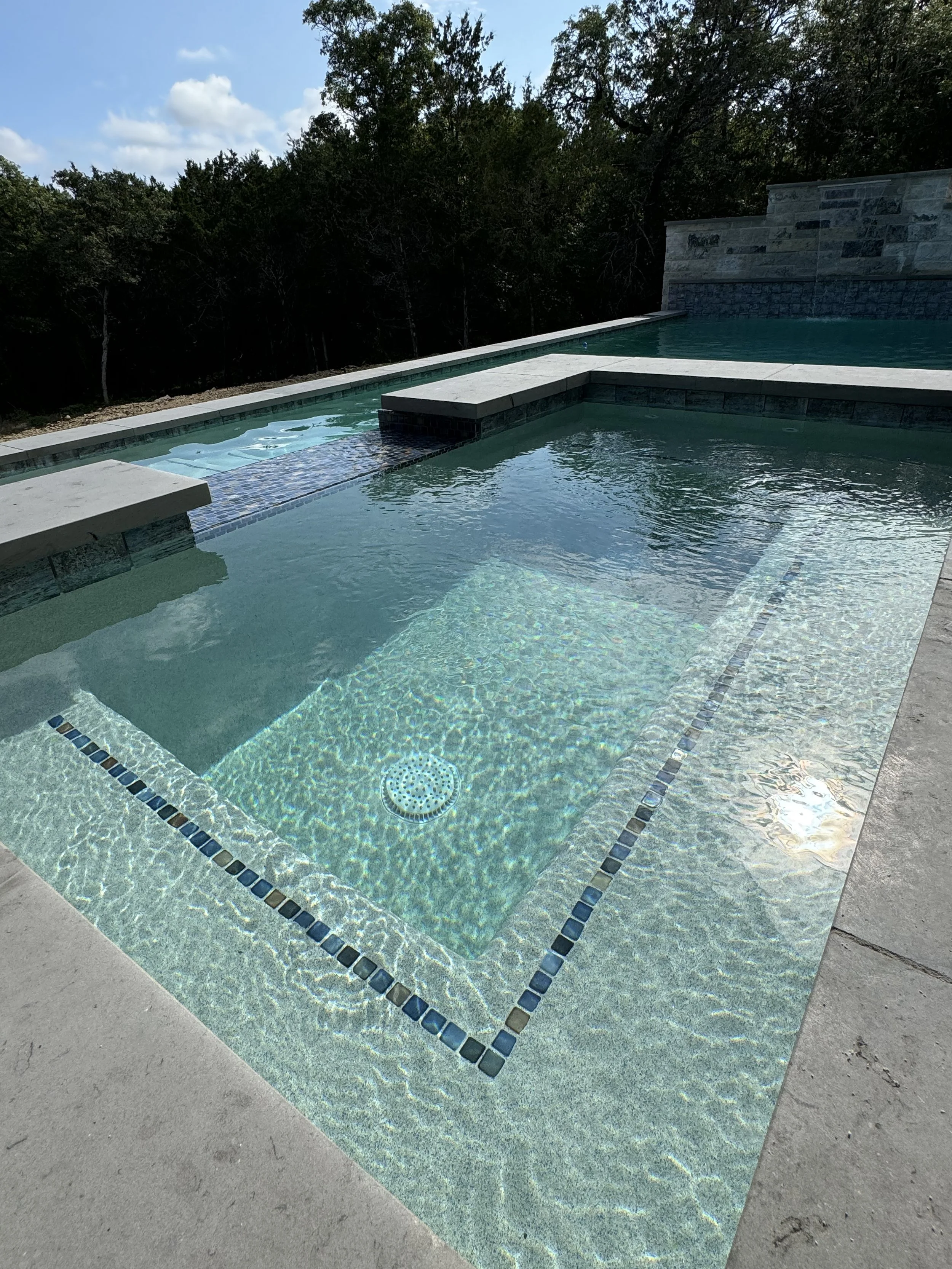 A modern in-ground swimming pool with a shallow area, surrounded by a concrete deck, with trees and a partly cloudy sky in the background.