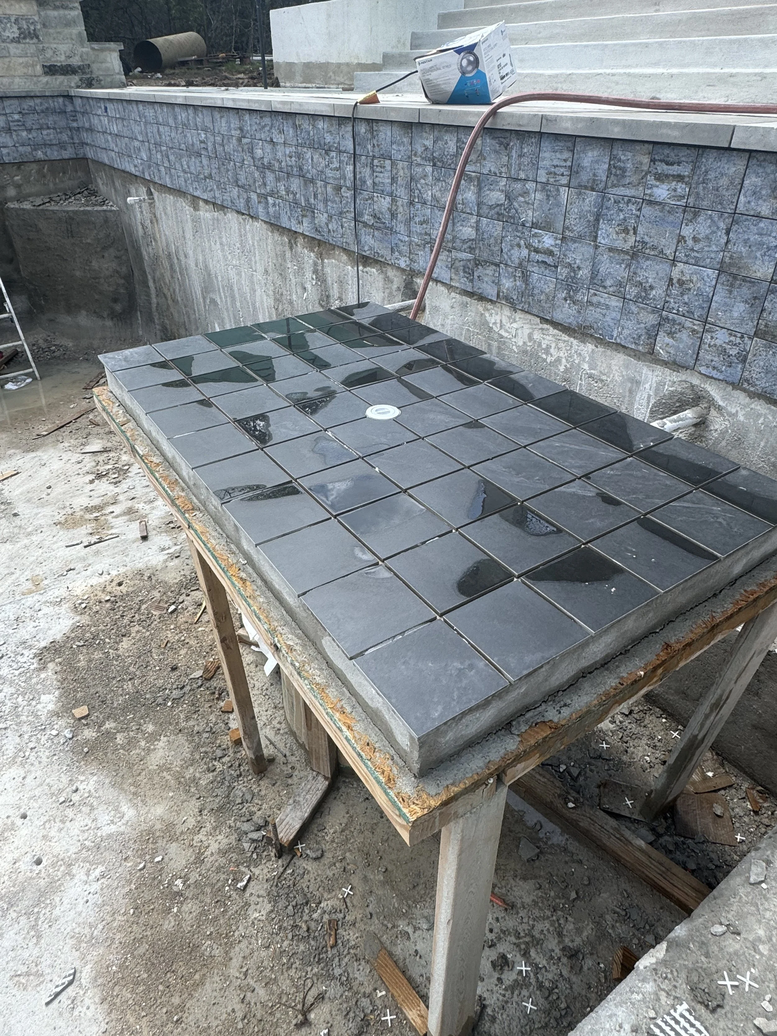 Construction site with a partially installed black tiled surface on a wooden worktable, surrounded by building materials, tools, and concrete steps.