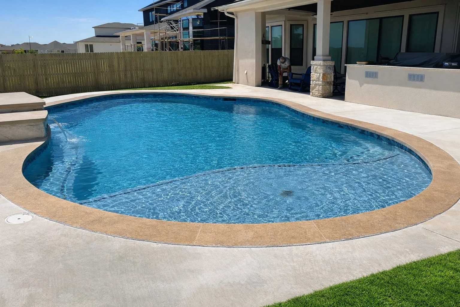 Backyard swimming pool with spa area and a covered patio, a man sitting on a chair, neighboring houses, wooden fence, green grass, clear blue sky.