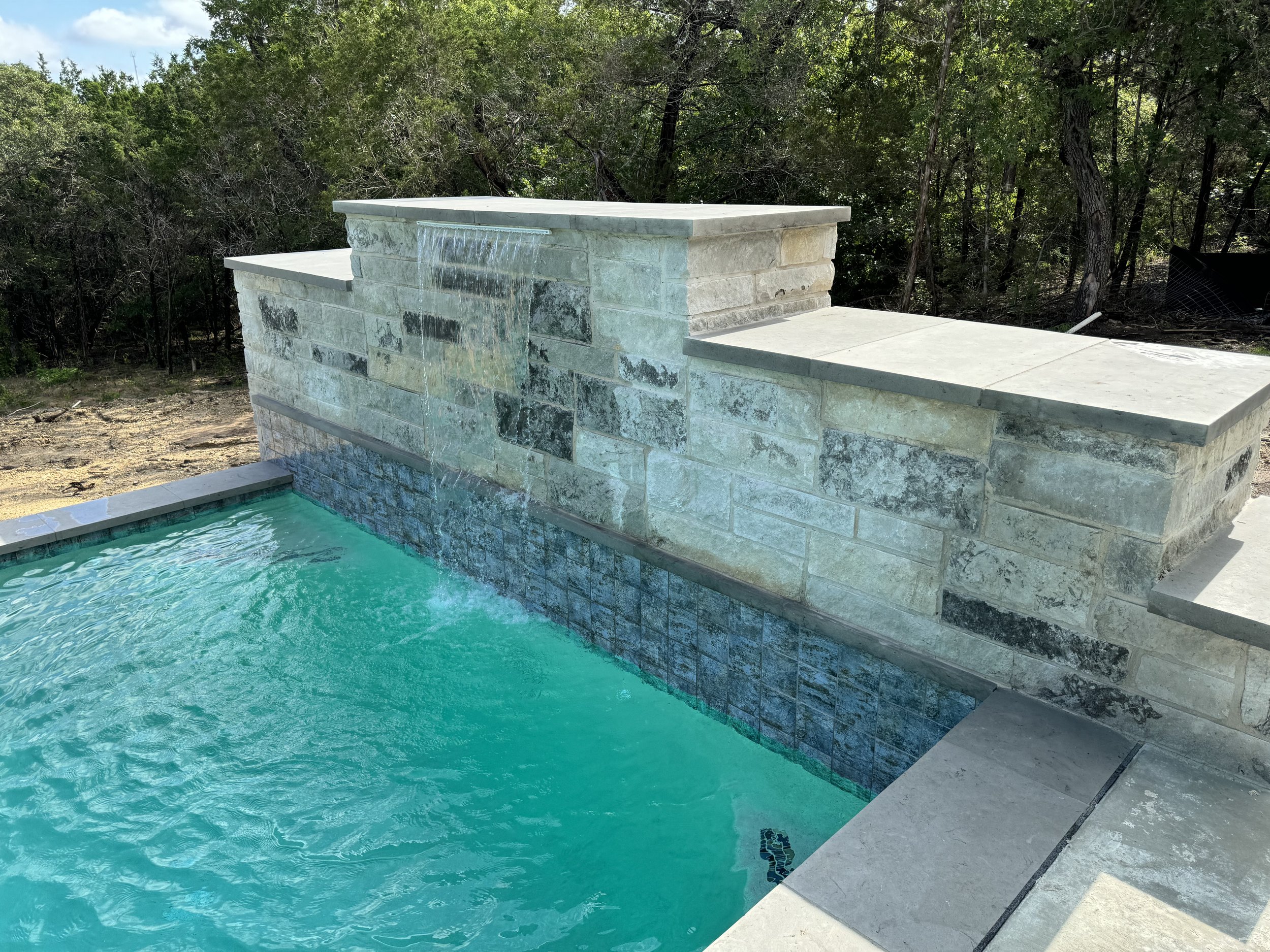 Above-ground swimming pool with a water feature flowing over a stone wall, surrounded by trees and outdoor patio stone flooring.