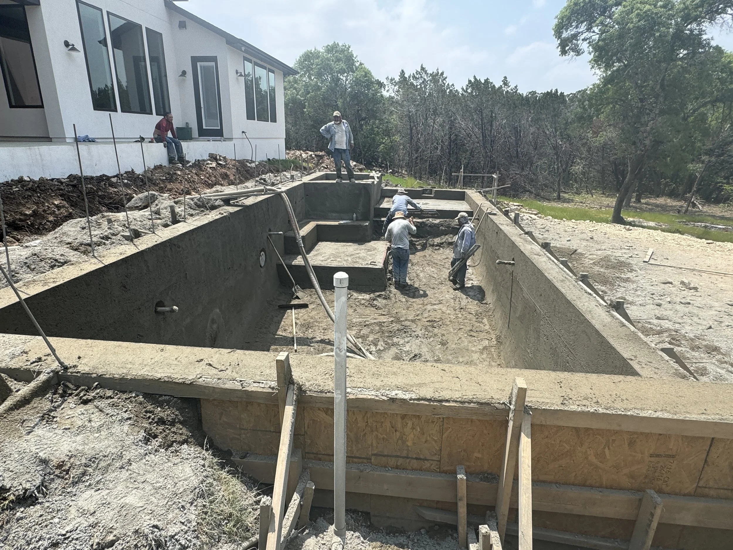 Construction workers building a swimming pool in a backyard, with a modern house to the left and trees in the background.