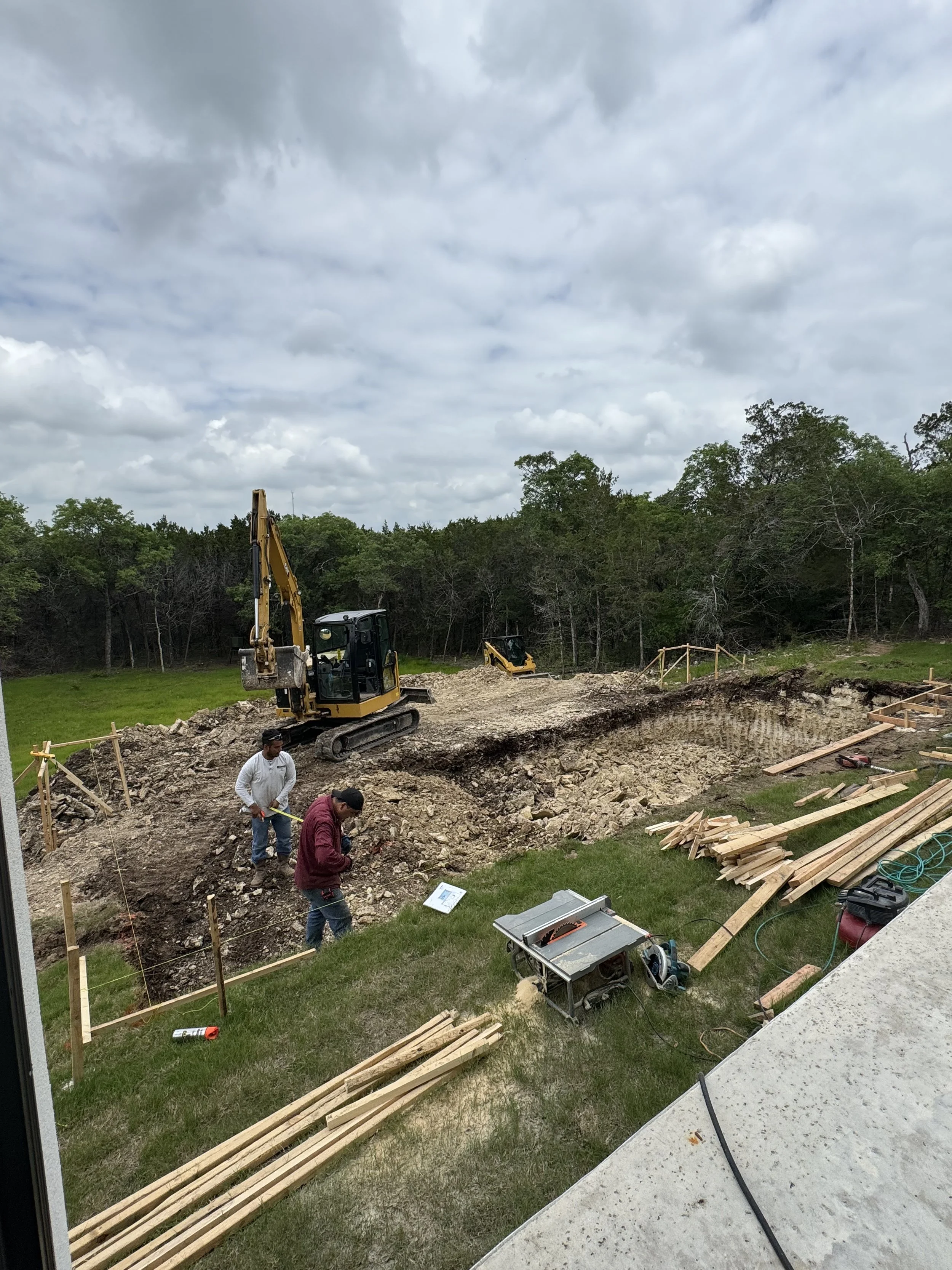 Construction workers and equipment working on a construction site outdoors, with an excavator moving dirt and wood planks organized nearby, under a cloudy sky.
