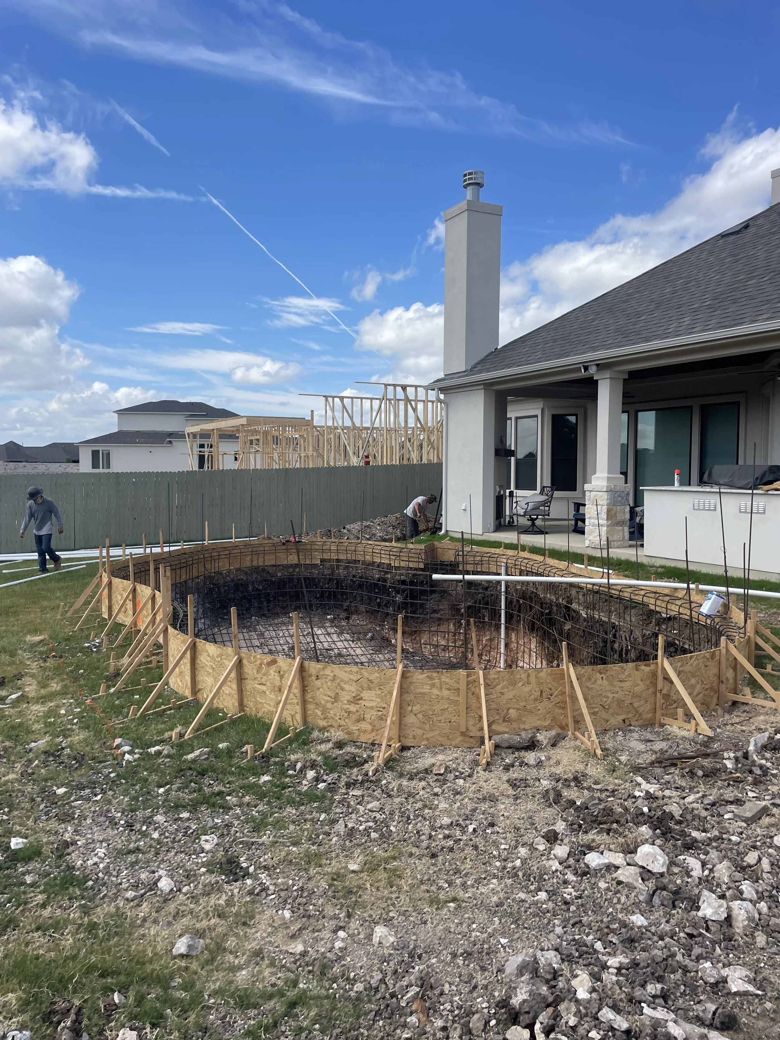 Backyard under construction with a dug hole for a swimming pool, wooden framing around the excavation, and construction workers working on the site.
