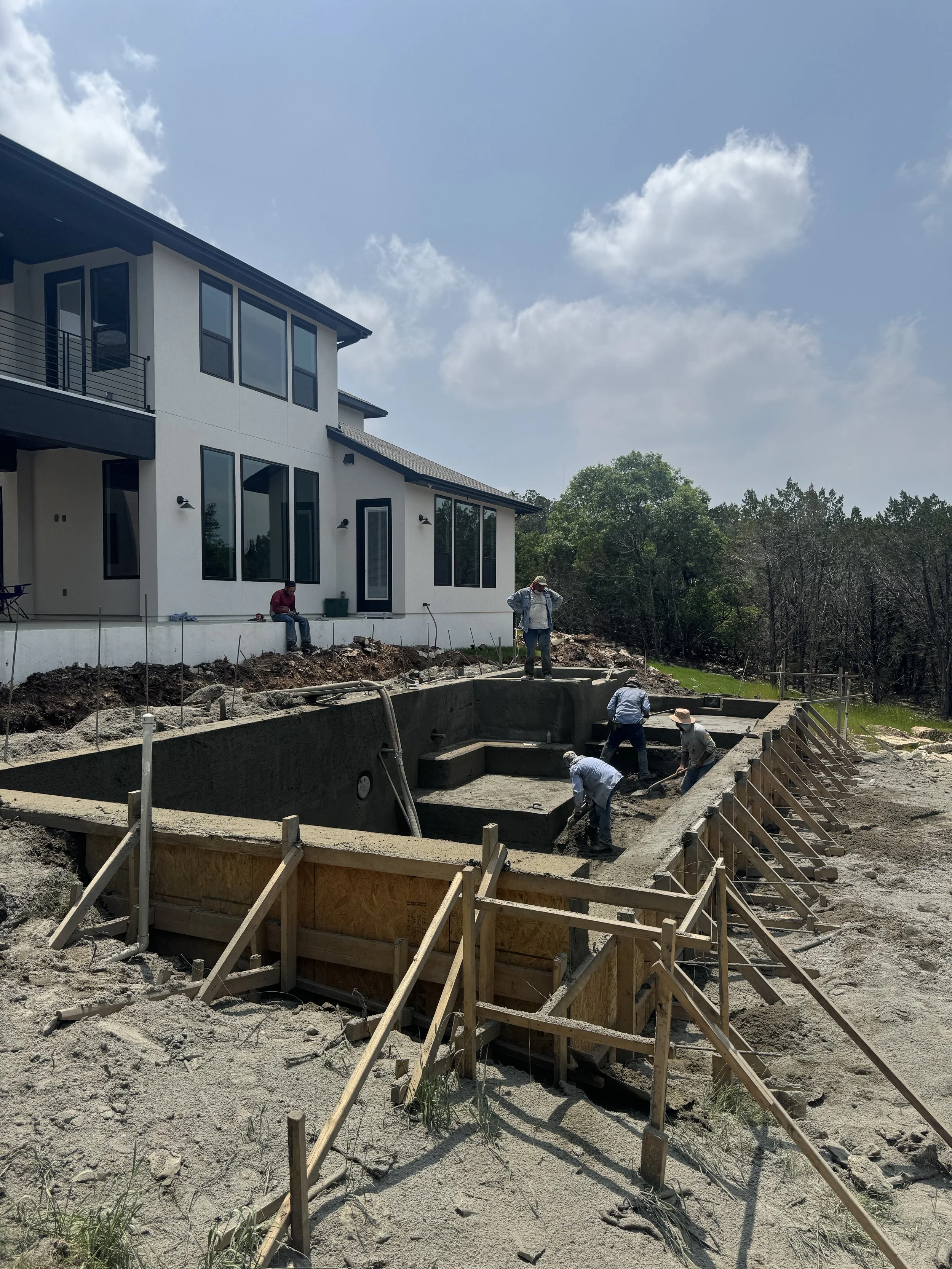 Construction workers building a swimming pool in the backyard of a modern white house on a sunny day with blue sky and clouds.