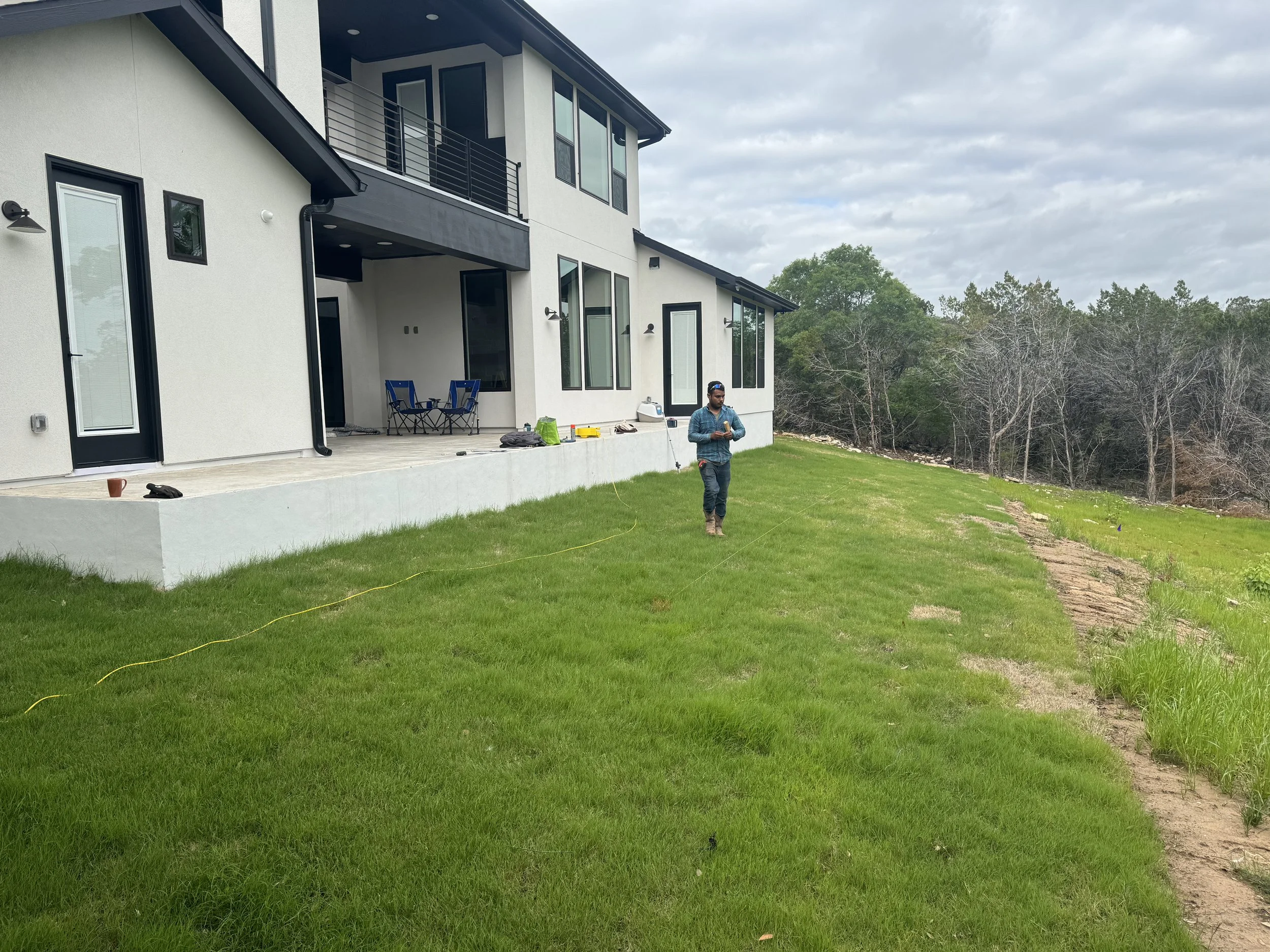 A man standing on a grassy backyard near a modern two-story house with a patio, under a cloudy sky, with trees in the background.