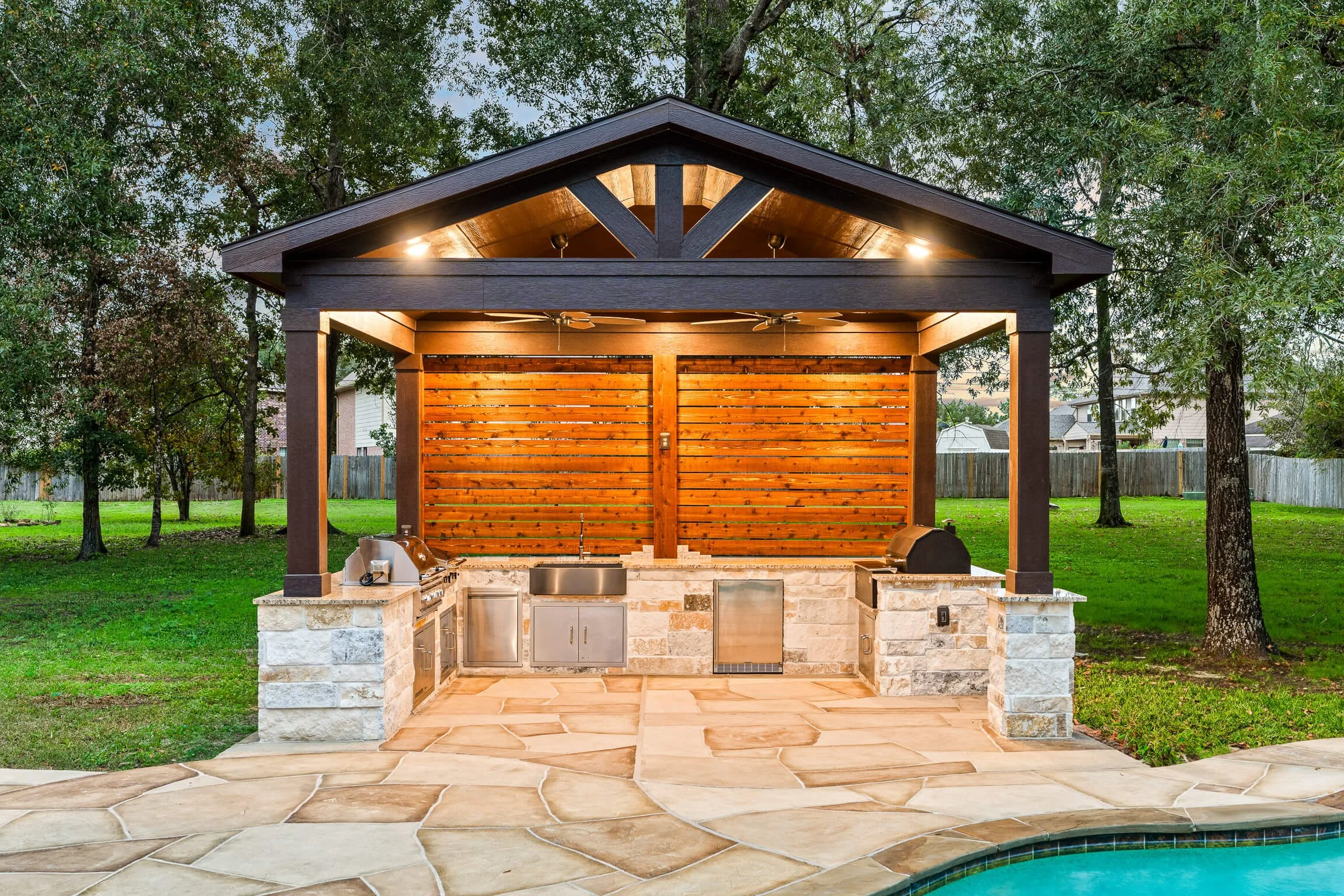 An outdoor kitchen with a stone counter, stainless steel appliances, and a wooden back wall under a roof with ceiling fans, surrounded by a grassy yard with trees.