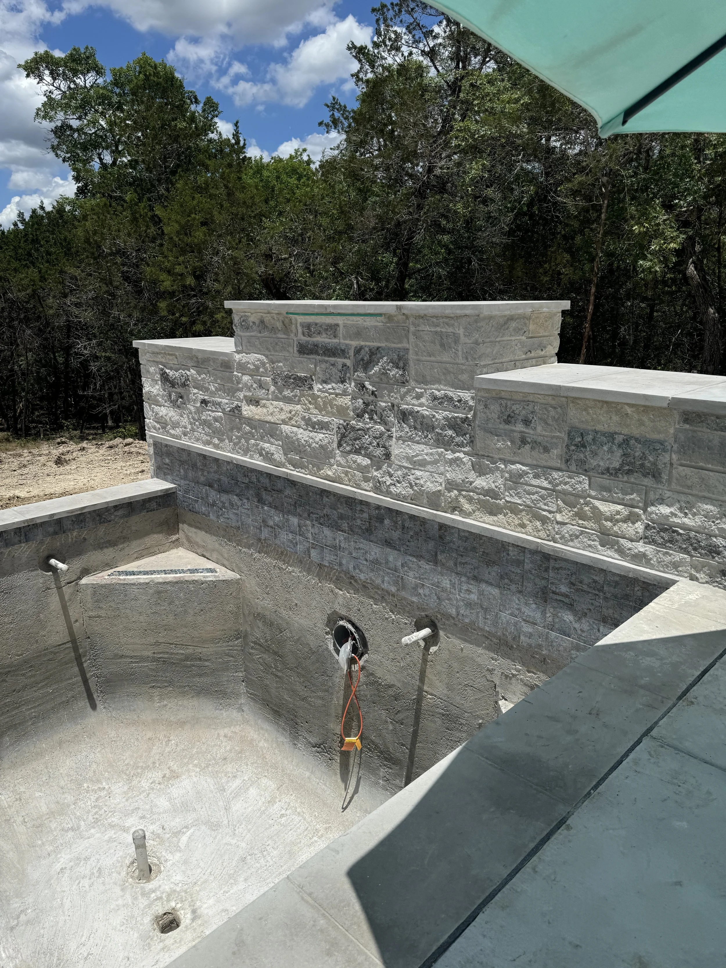 An outdoor swimming pool under construction with a concrete basin, surrounded by a stone and brick wall, trees in the background, and a blue sky with clouds.