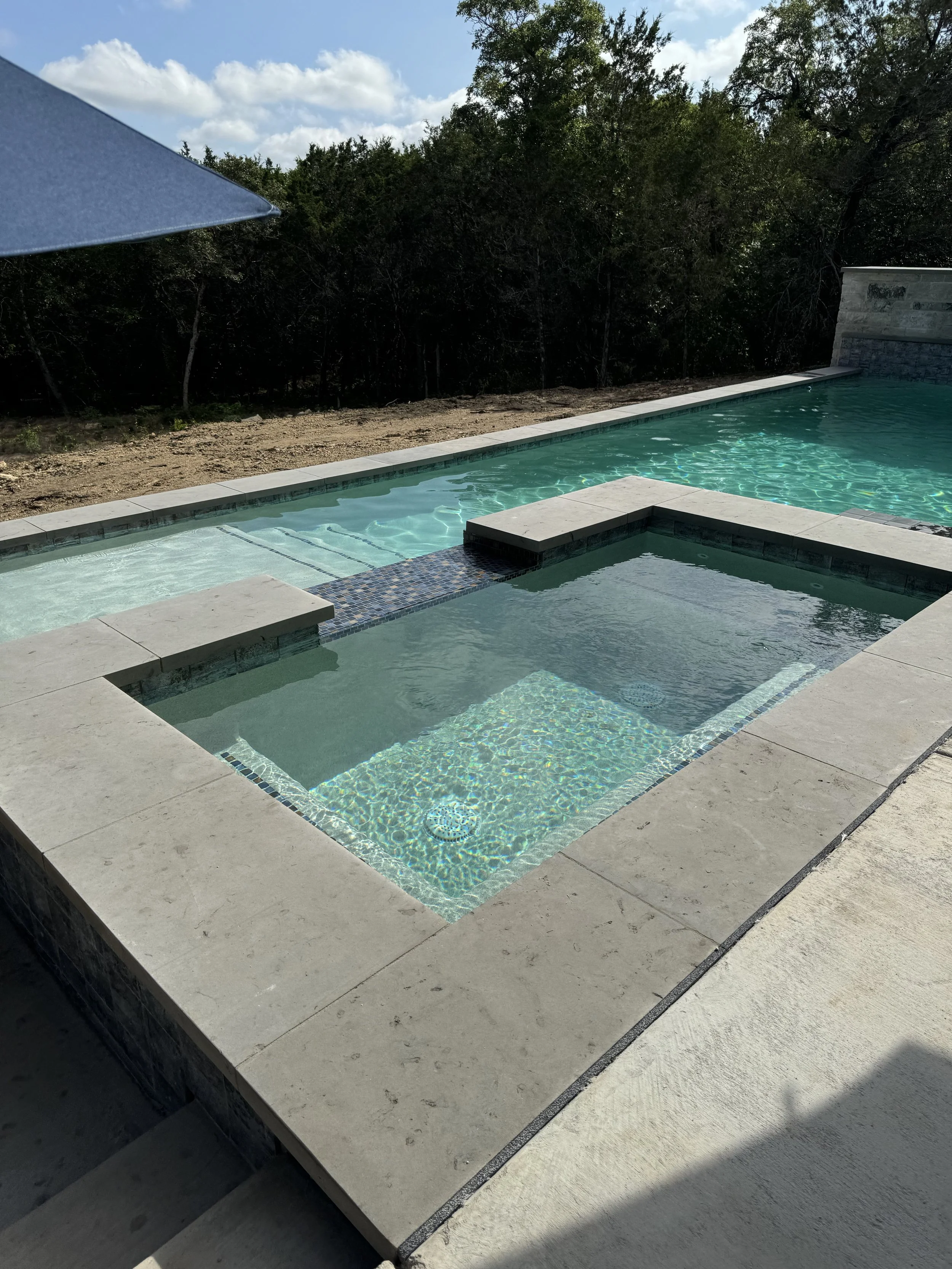 A backyard modern swimming pool with a connected hot tub, beige stone coping, a blue patio umbrella on the left, and a row of trees in the background, under a partly cloudy sky.