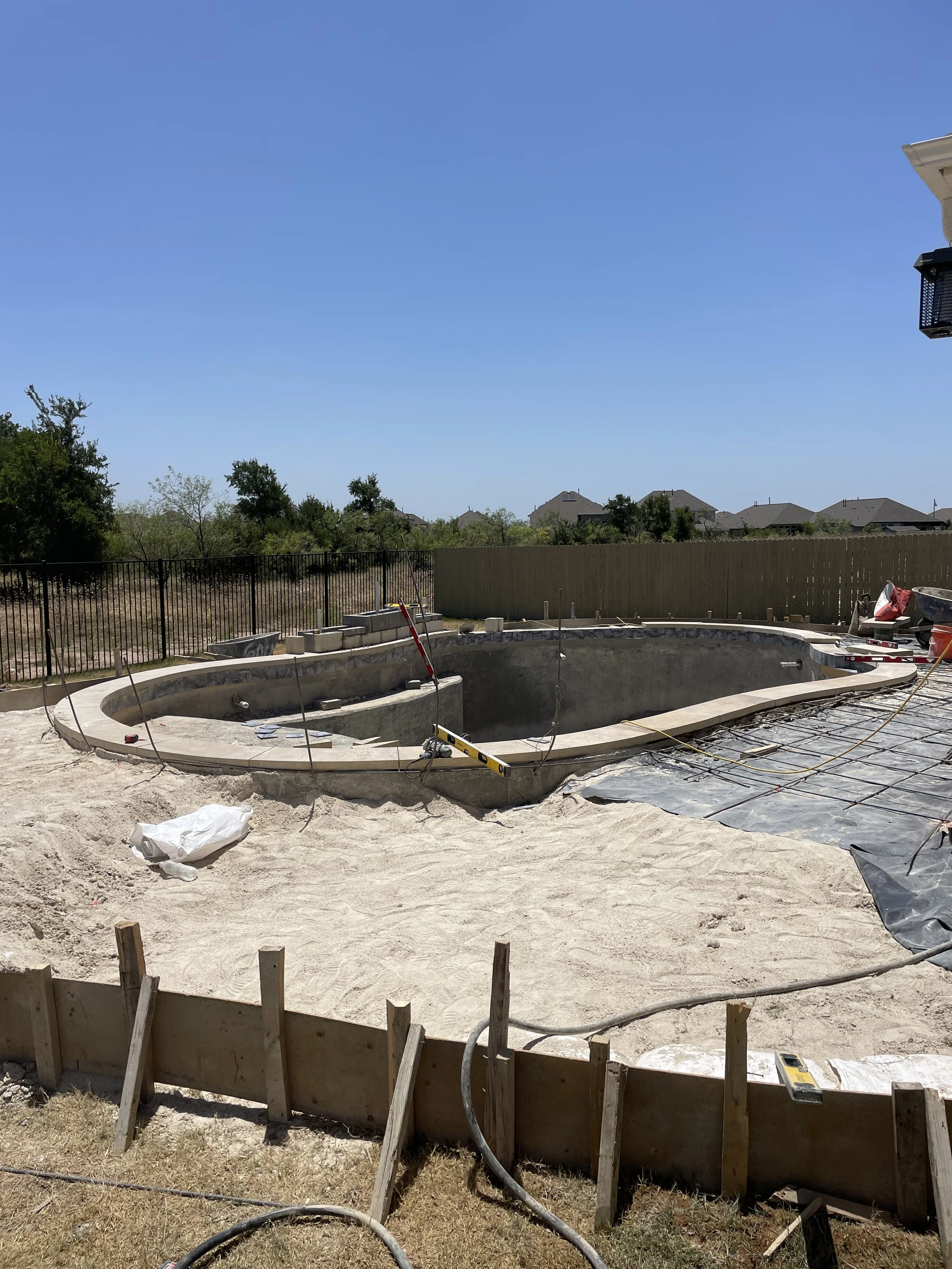 Under construction backyard swimming pool with curved concrete edge, surrounded by construction materials, fencing, and tools, with a clear blue sky overhead.