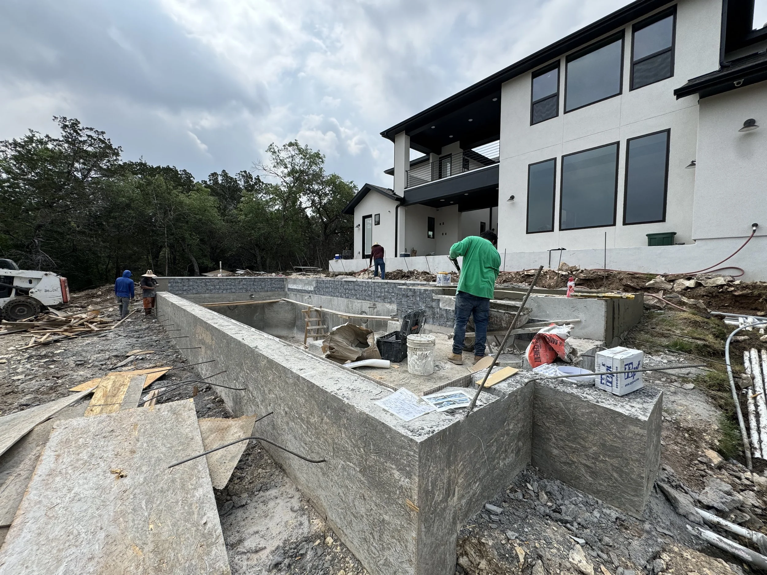 Construction workers building a concrete pool in the backyard of a modern house under cloudy skies.