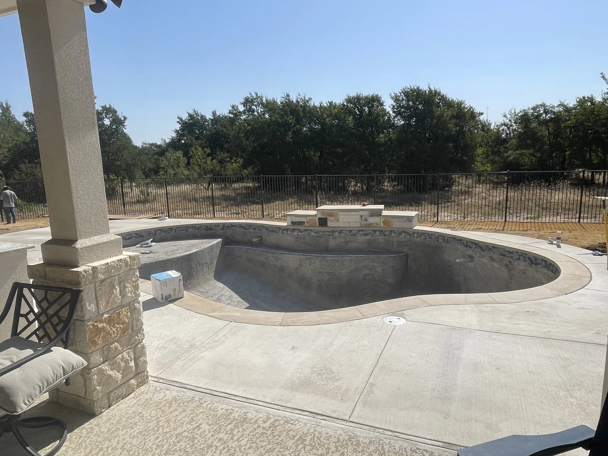 Empty in-ground swimming pool under construction with a curved shape, surrounded by a concrete deck, and a stone bench at the far end. Fencing and trees are visible in the background.