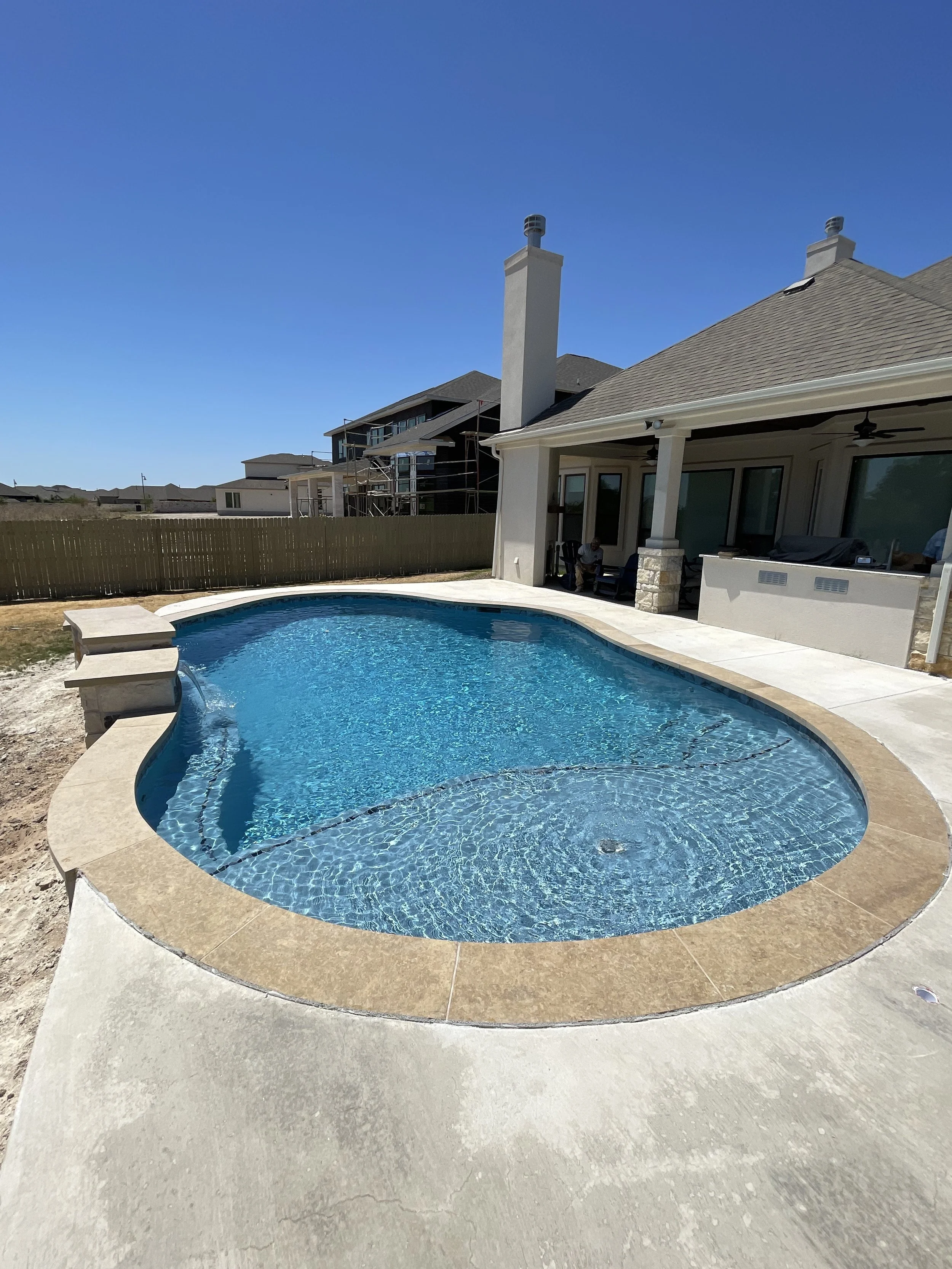 Backyard with an in-ground swimming pool, patio, and house with a chimney, under a clear blue sky.