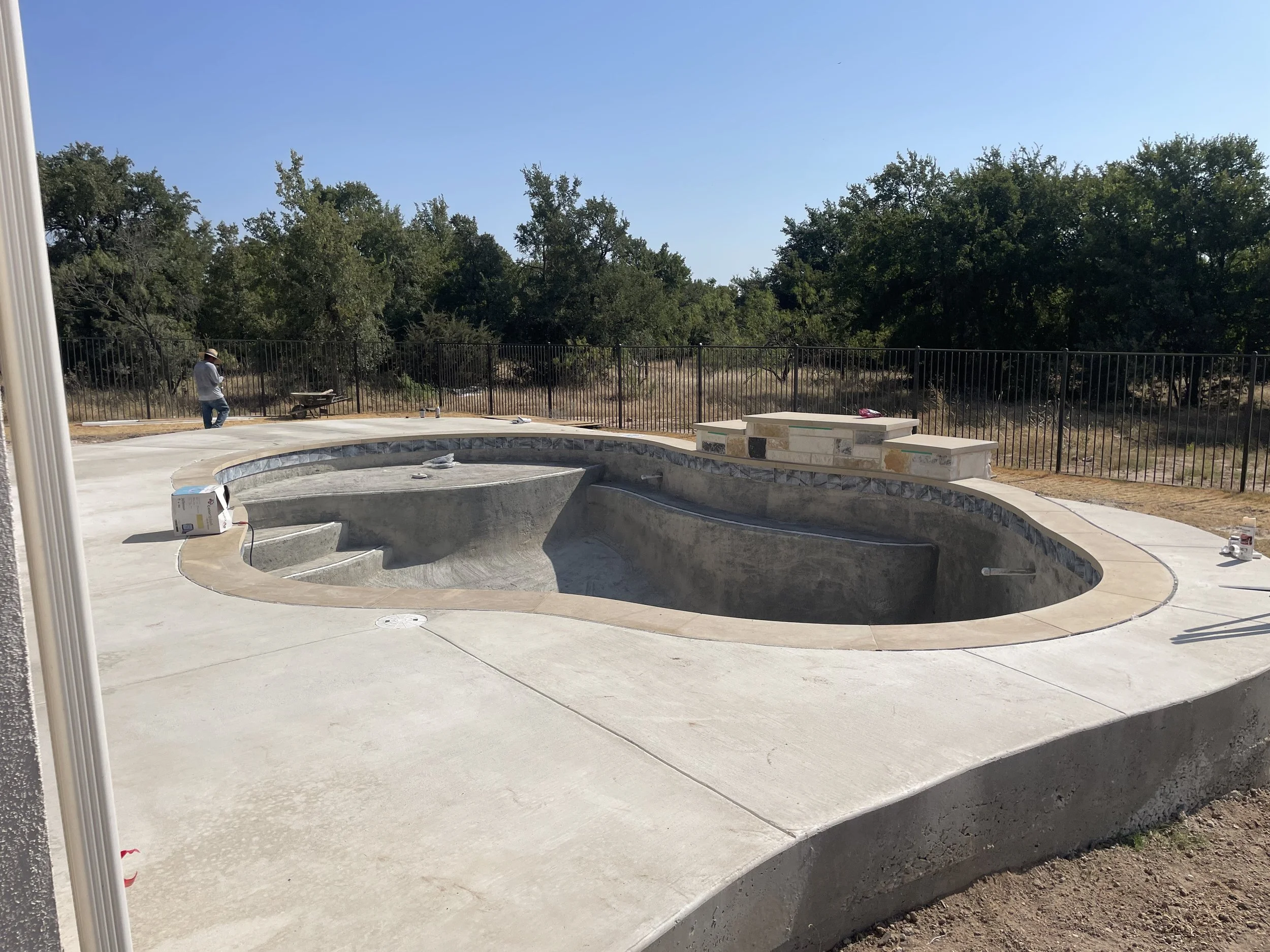 An under-construction backyard swimming pool with a kidney shape, surrounded by a concrete deck and a decorative stone border. A worker is visible in the background near a fence, and construction tools are scattered on the deck.