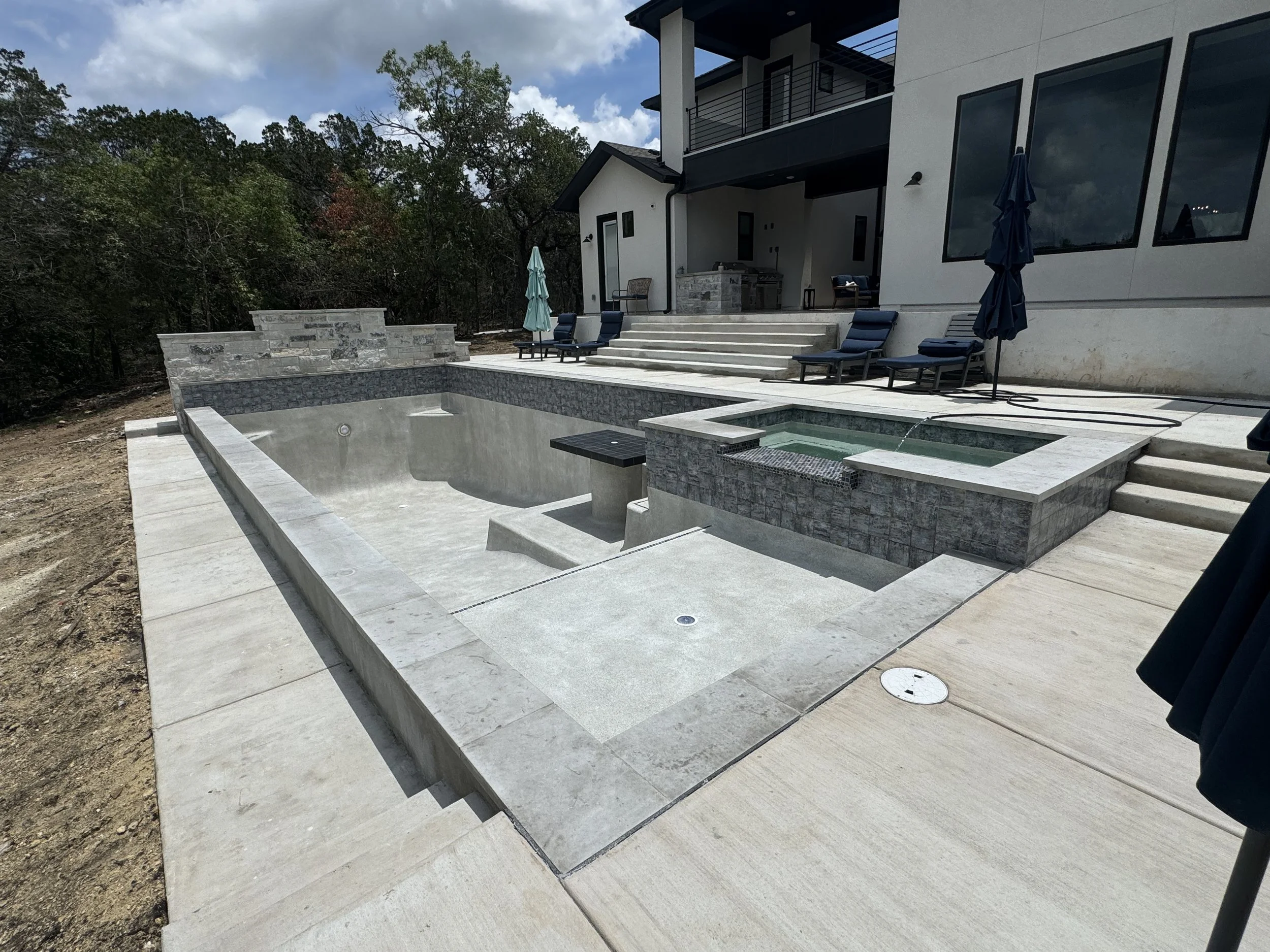 Modern backyard with a large, empty in-ground swimming pool, a connected hot tub, and neighboring patio furniture with umbrellas, outside a contemporary house with large windows and stairs leading up to the house.