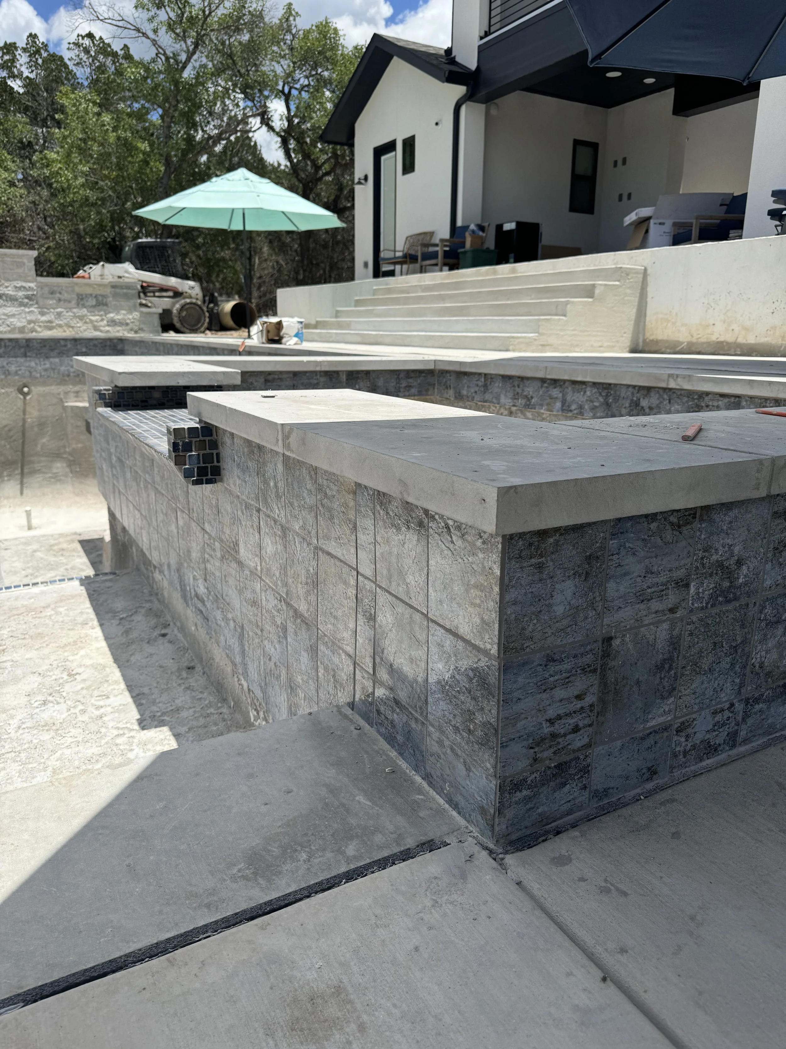 Construction site of an outdoor pool with tile and concrete finishing, steps leading up to a house with chairs, umbrella, and construction materials in the background.