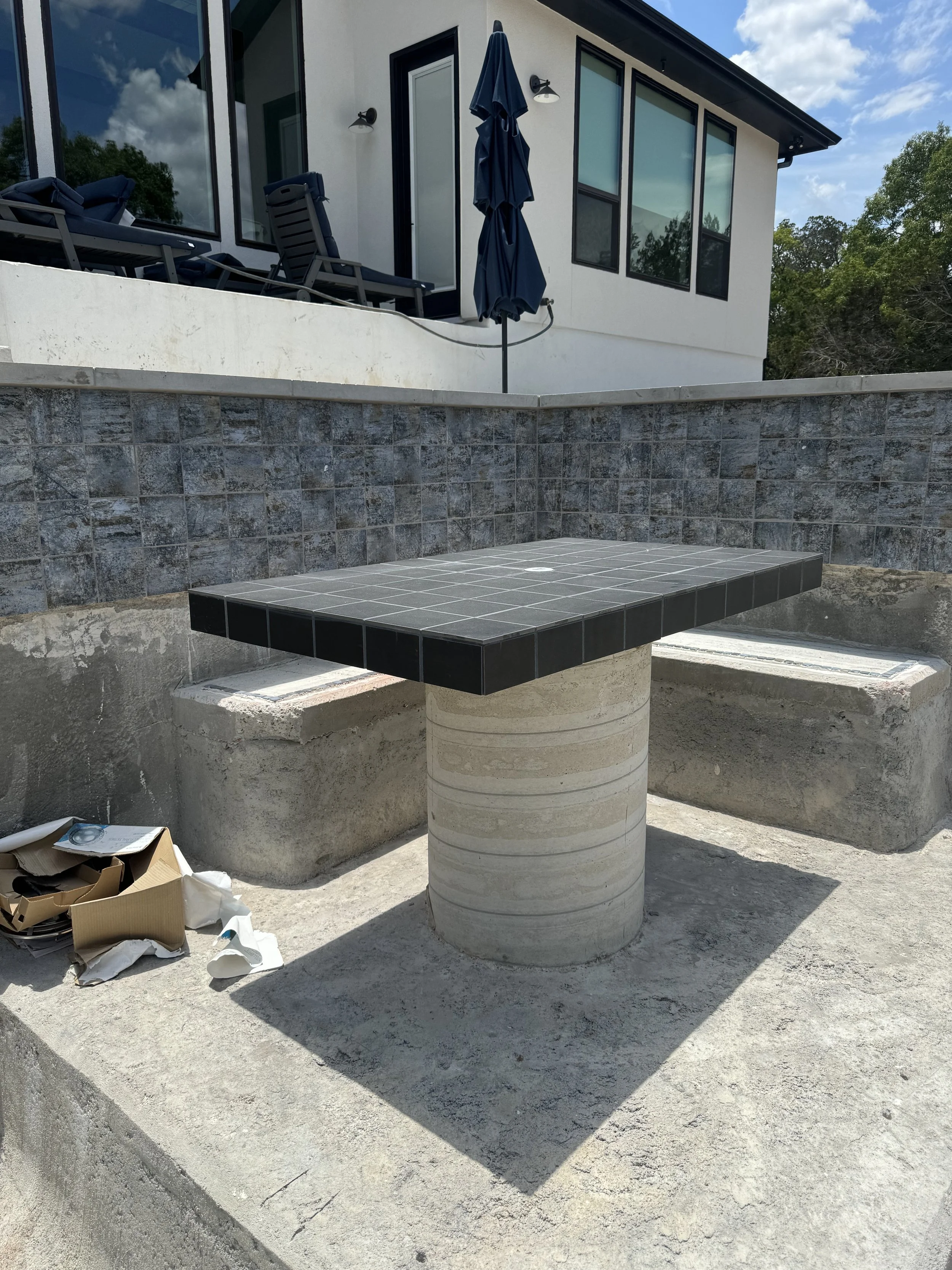 A modern outdoor table with a tiled black top and concrete cylindrical base, situated on a concrete patio against a retaining wall and overlooking a house with patio chairs and a closed umbrella.